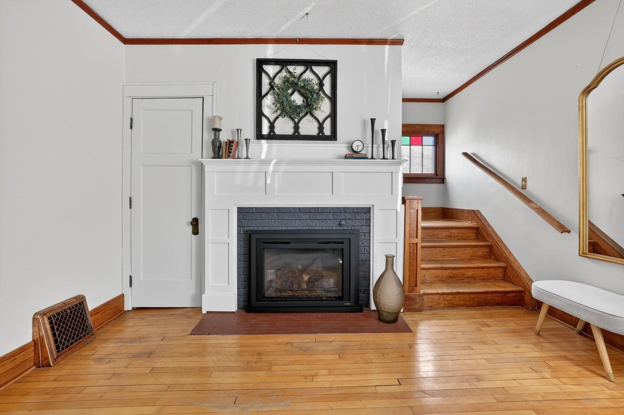 Living room with a fireplace, wooden stairs, and decor on the mantel, including a wreath, books, candles, and vases, with hardwood flooring and a window with colorful stained glass.