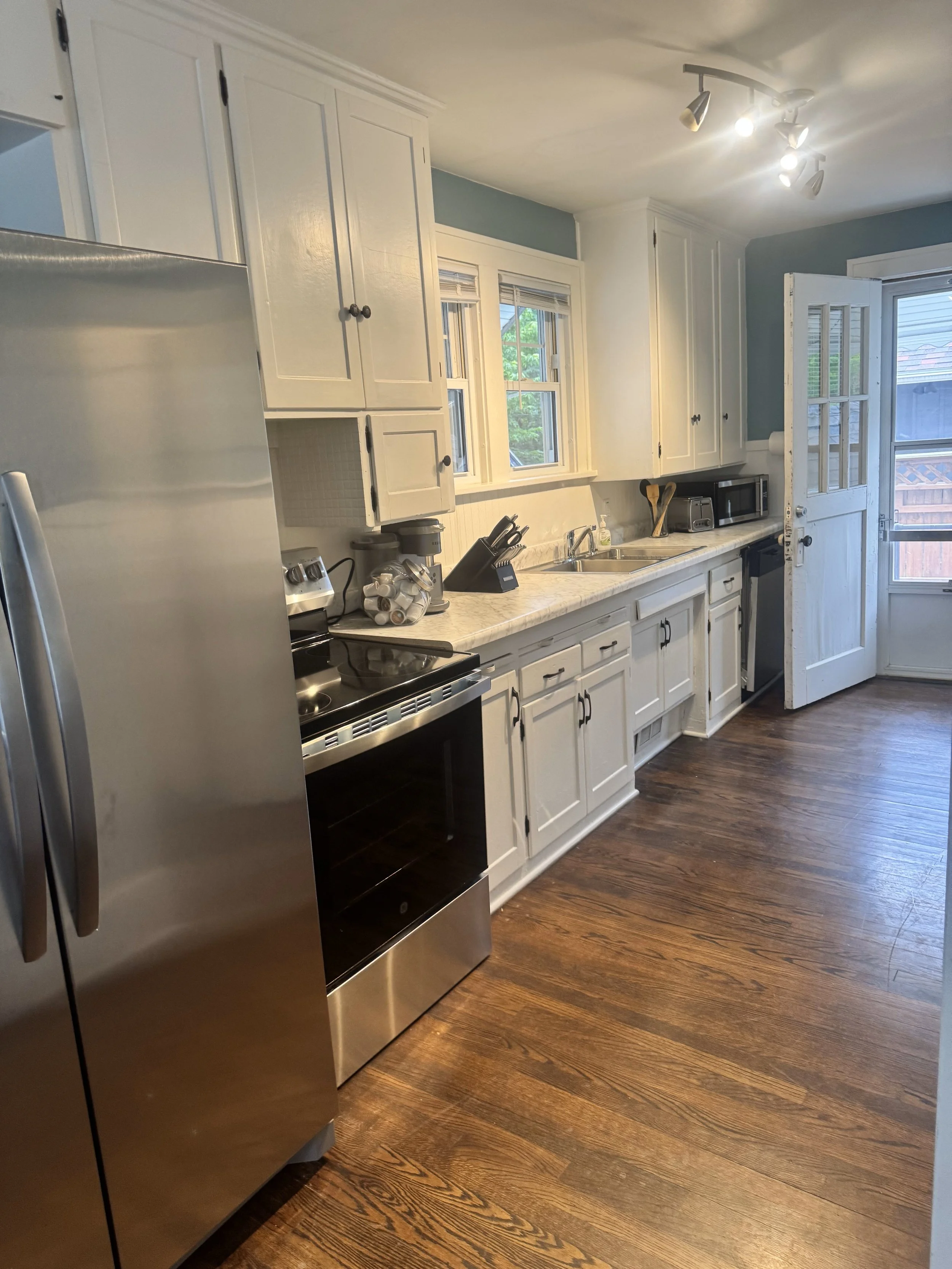 Kitchen with white cabinets, stainless steel refrigerator, microwave, black stovetop, wooden floor, and a door leading outside.