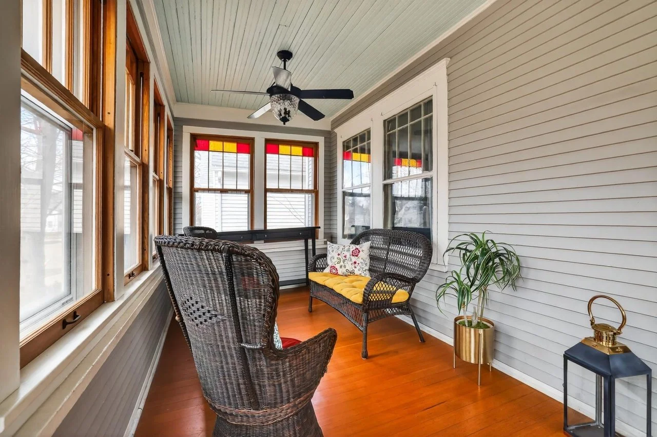 Sunroom with wicker chairs, a yellow cushion, a pillow, a potted plant, and a ceiling fan with a decorative light, featuring windows with stained glass accents.