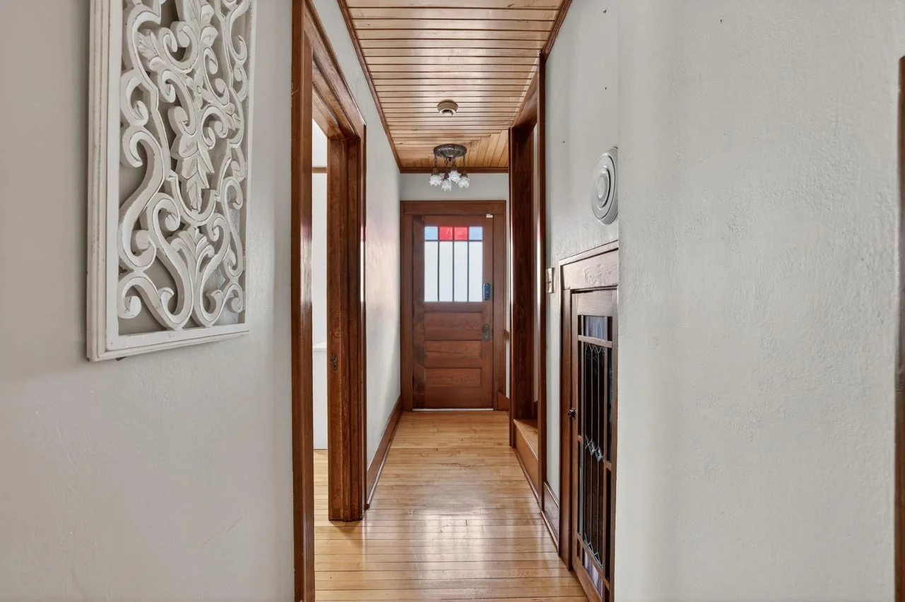 A narrow hallway with wooden floors, white walls, and wooden trim. There is a decorative white wall hanging on the left, a wooden door at the end with colored glass, and a small door with a metal grill on the right.