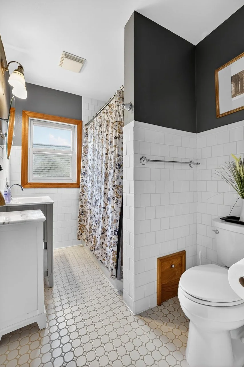 A small bathroom with white hexagonal tiled floor, white tiled walls, and dark gray walls above. It has a wooden framed window, a floral shower curtain, a white vanity, and a toilet with a decorative plant on top. There's a framed picture on the wall and a towel bar on the wall near the shower.