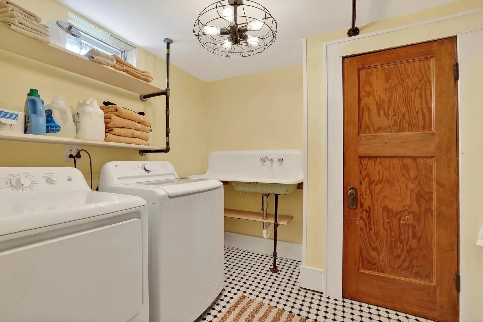 Laundromat laundry room with washing machine, utility sink, wooden door, wall shelves with towels and cleaning supplies, decorative ceiling light, black and white tiled floor.