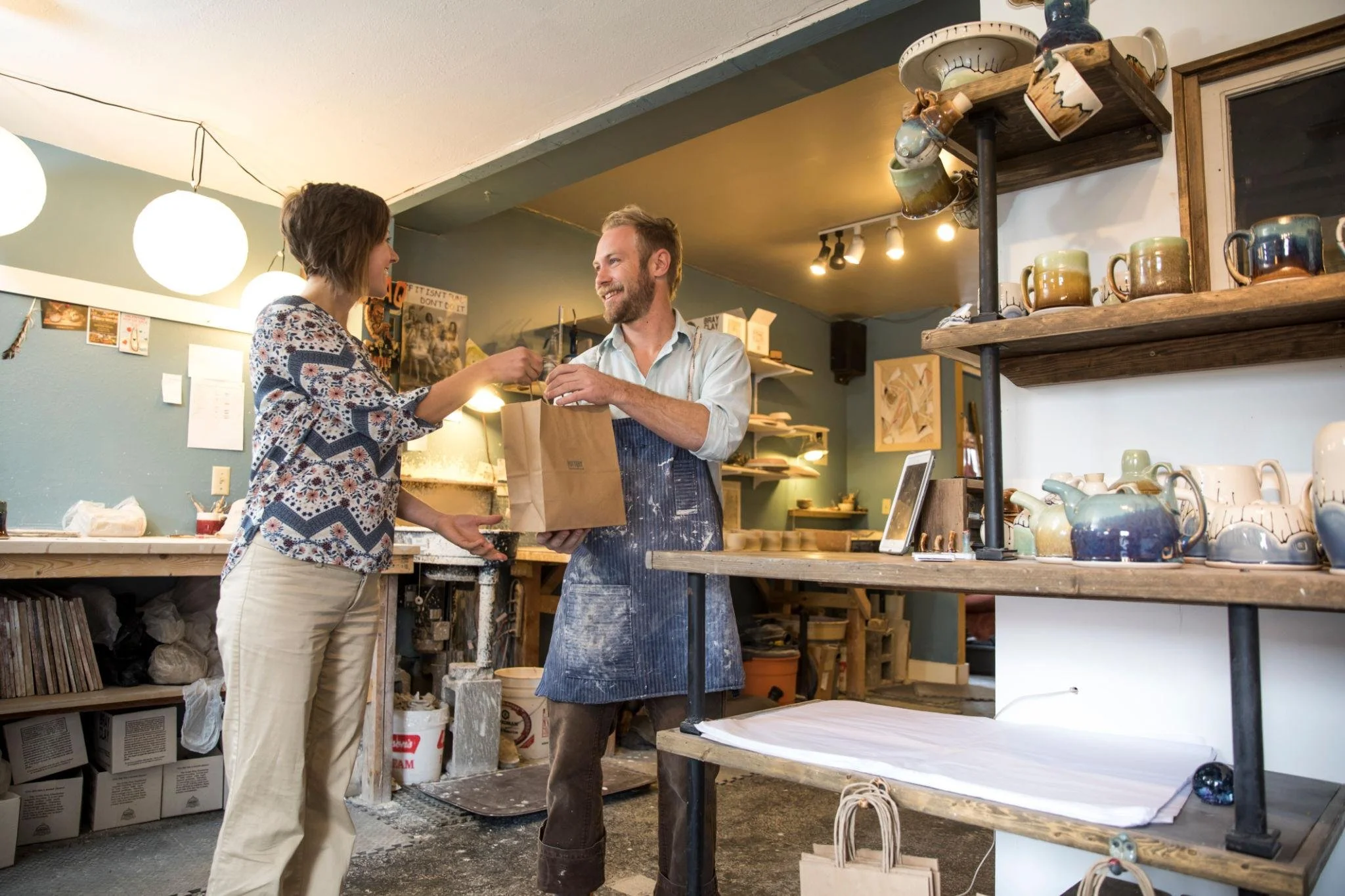 A woman and a man exchanging a paper bag in a ceramic studio. The woman has short brown hair and is wearing a patterned blouse and beige pants. The man has a beard, short hair, and is wearing a light shirt with an apron. They are standing near shelve