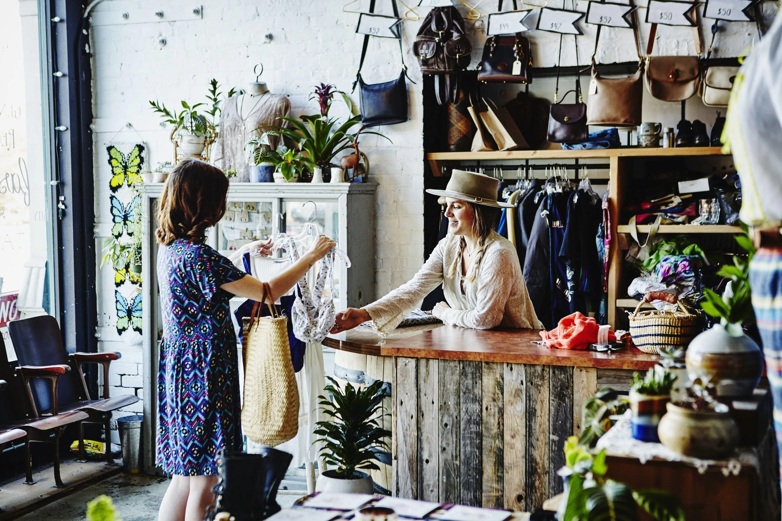 Two women are shopping for clothes inside a boutique store. One woman with long hair and a colorful dress is holding a yellow dress, while the other woman, wearing a beige hat and a cream-colored lace top, is sitting behind a wooden counter smiling. 