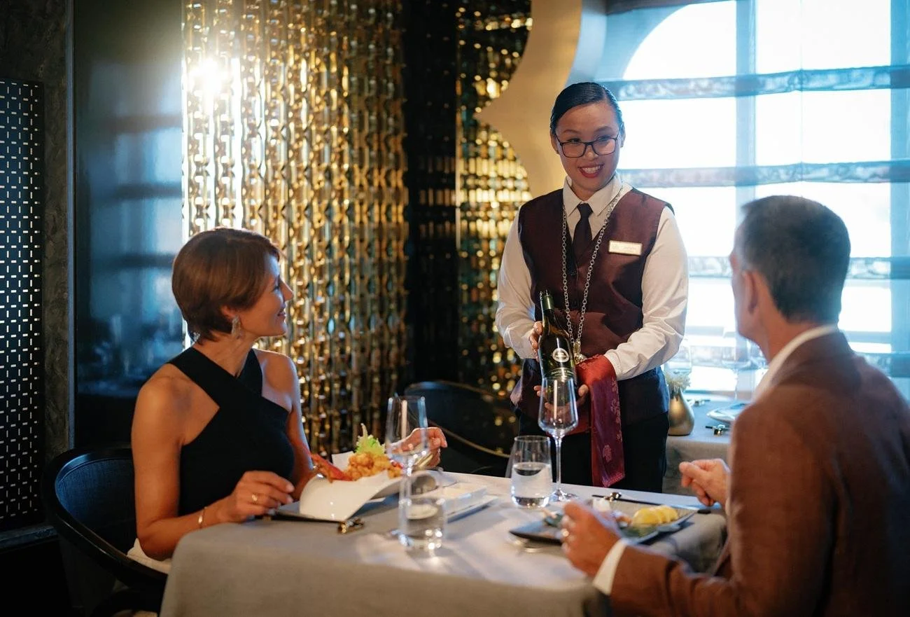 A woman in a black dress eating dinner at a restaurant table with a man in a tan suit. A waitress in a uniform serves them wine in a glass. The restaurant has modern decor and large windows with curtains.
