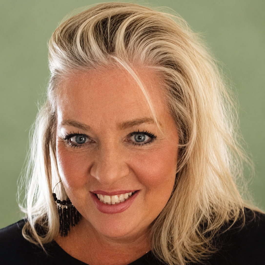 Close-up portrait of a smiling woman with blonde hair and blue eyes, wearing black earrings and a black top against a muted green background.