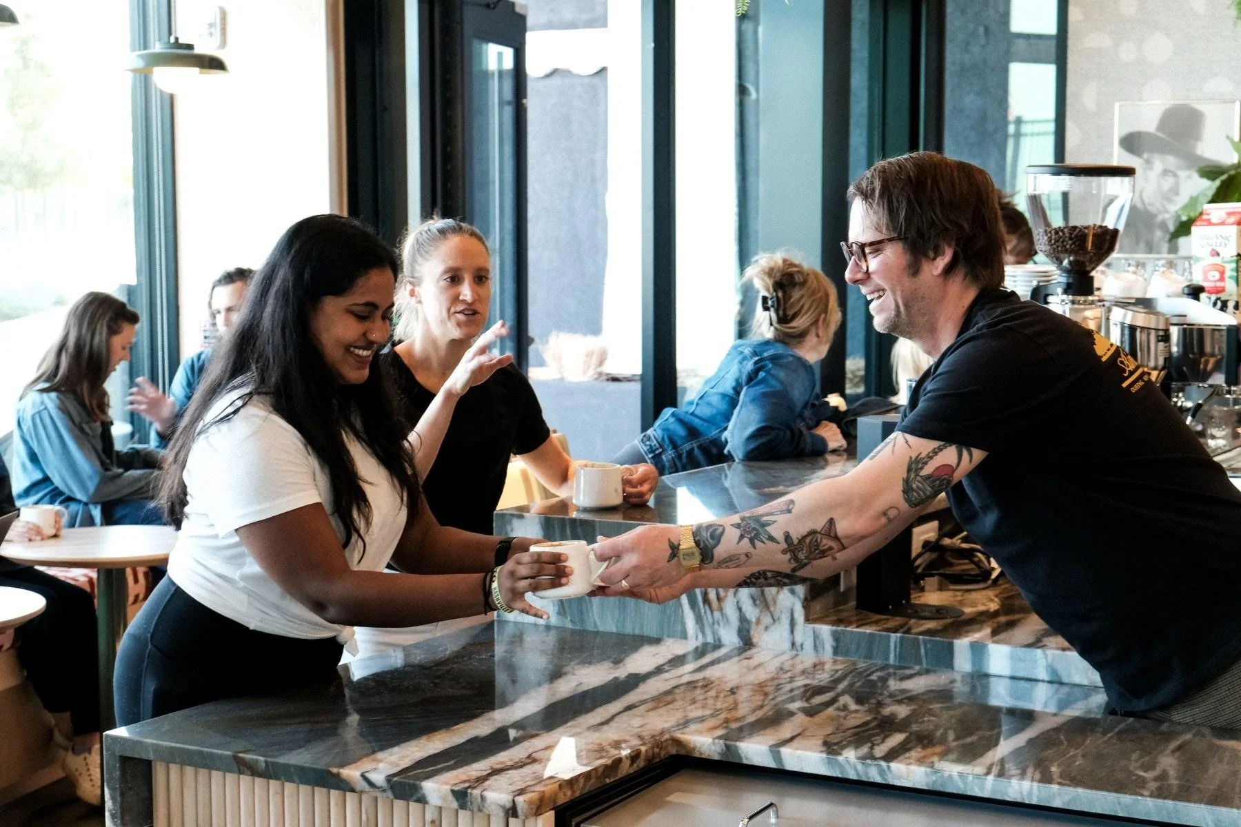Barista smiling and handing a coffee cup to a woman in a cafe. Several customers are sitting in the background by large windows.