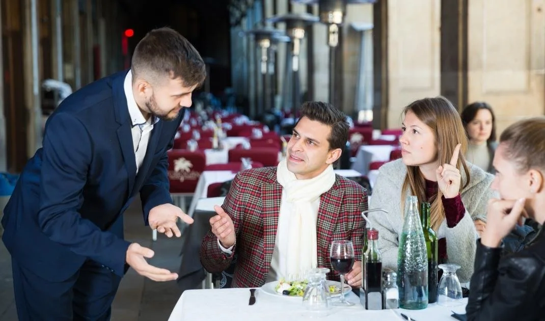Man in a blue suit gesturing while talking to a man in a brown plaid blazer at a restaurant table, with two women nearby, one holding a fork and looking attentive, and the other with her hand on her chin. The table has wine glasses and condiments.