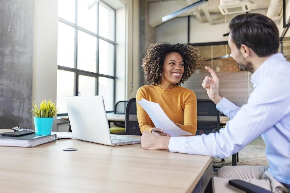 A woman and a man having a discussion in an office, with the woman smiling and holding papers, and the man gesturing with his finger.