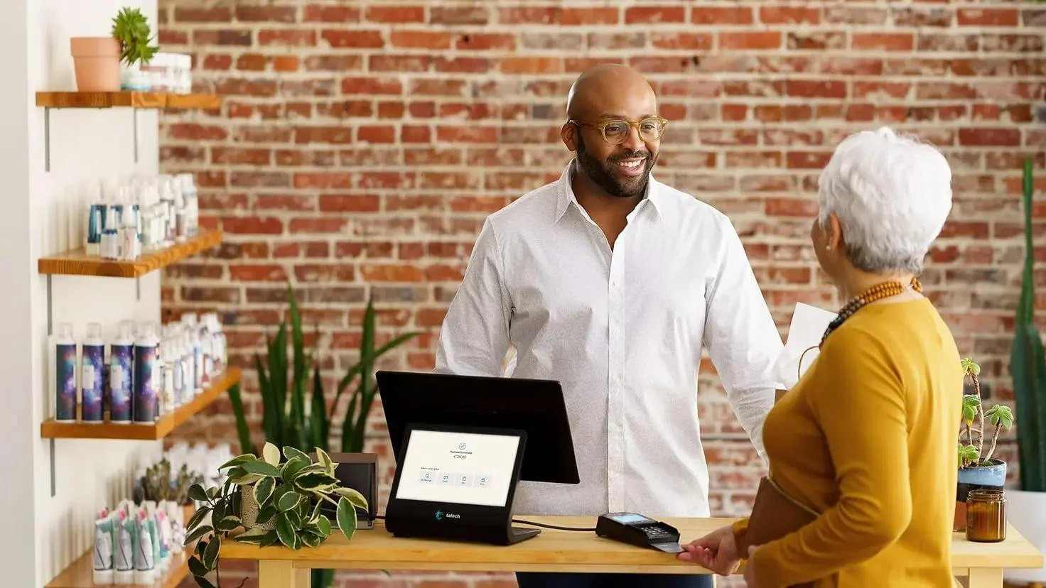 A man with glasses and a beard smiling at a woman with white hair in a yellow sweater. They are standing in a store or shop with a brick wall in the background, a computer on the counter, and shelves with bottles and plants.