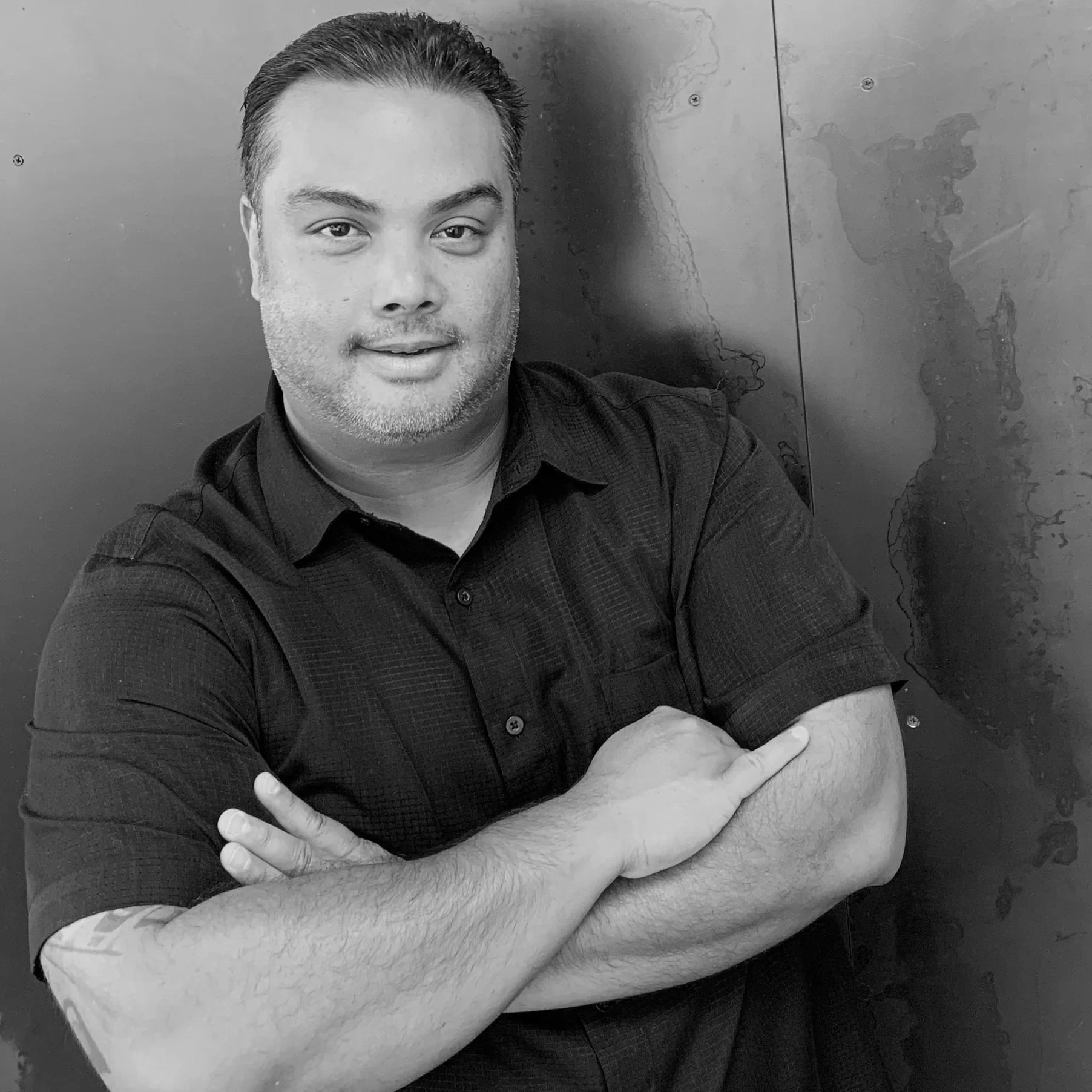 A man taking a selfie with his arms crossed, wearing a dark shirt, in front of a wall with a map outline.