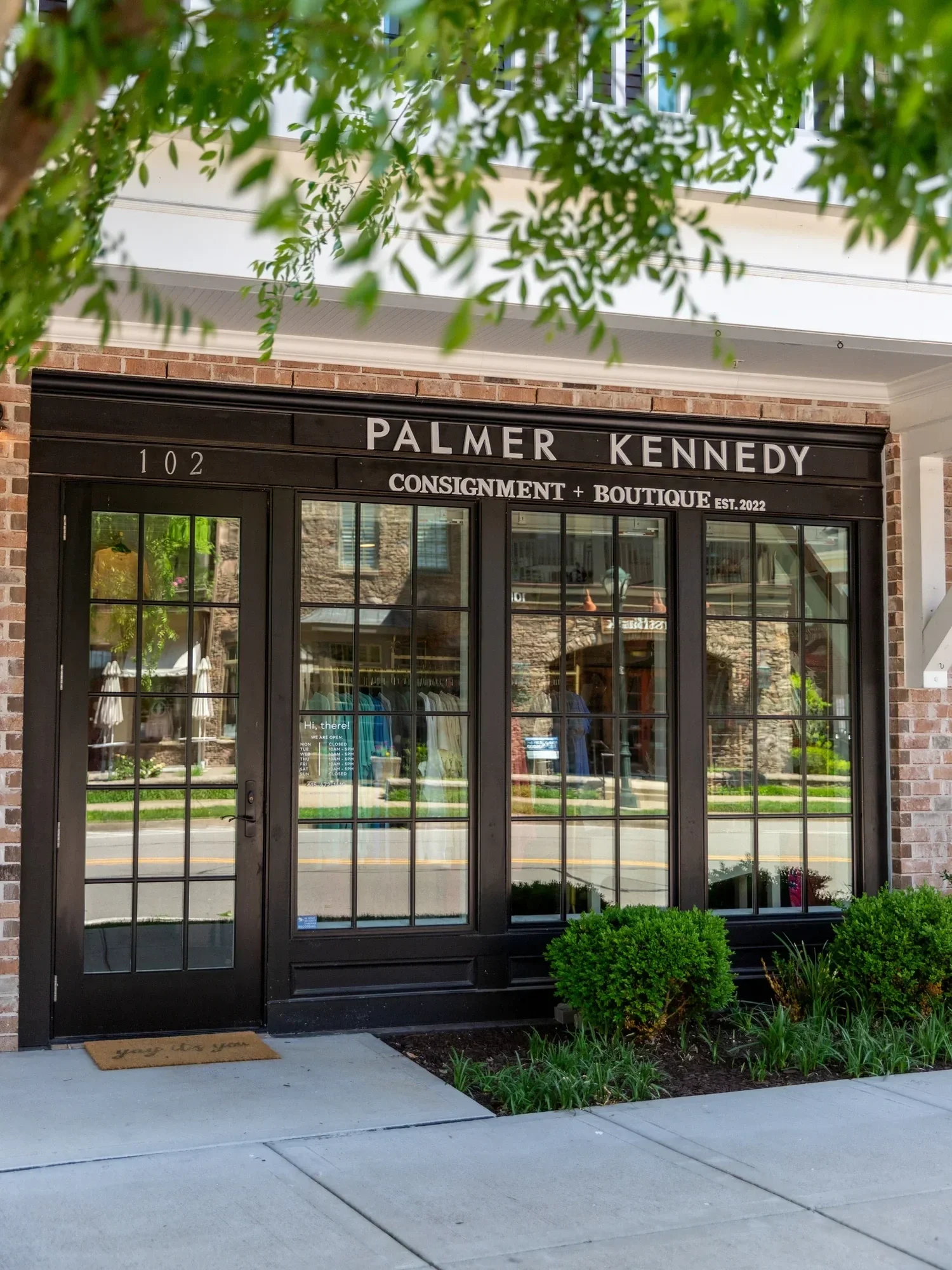 The storefront of Palmer Kennedy, a consignment and boutique shop, with the street reflected in the glass doors and window. The sign indicates the store was established in 2022 and is located at 102. There is a green shrub and some plants outside, and a doormat with the words 'hello to you' near the entrance.