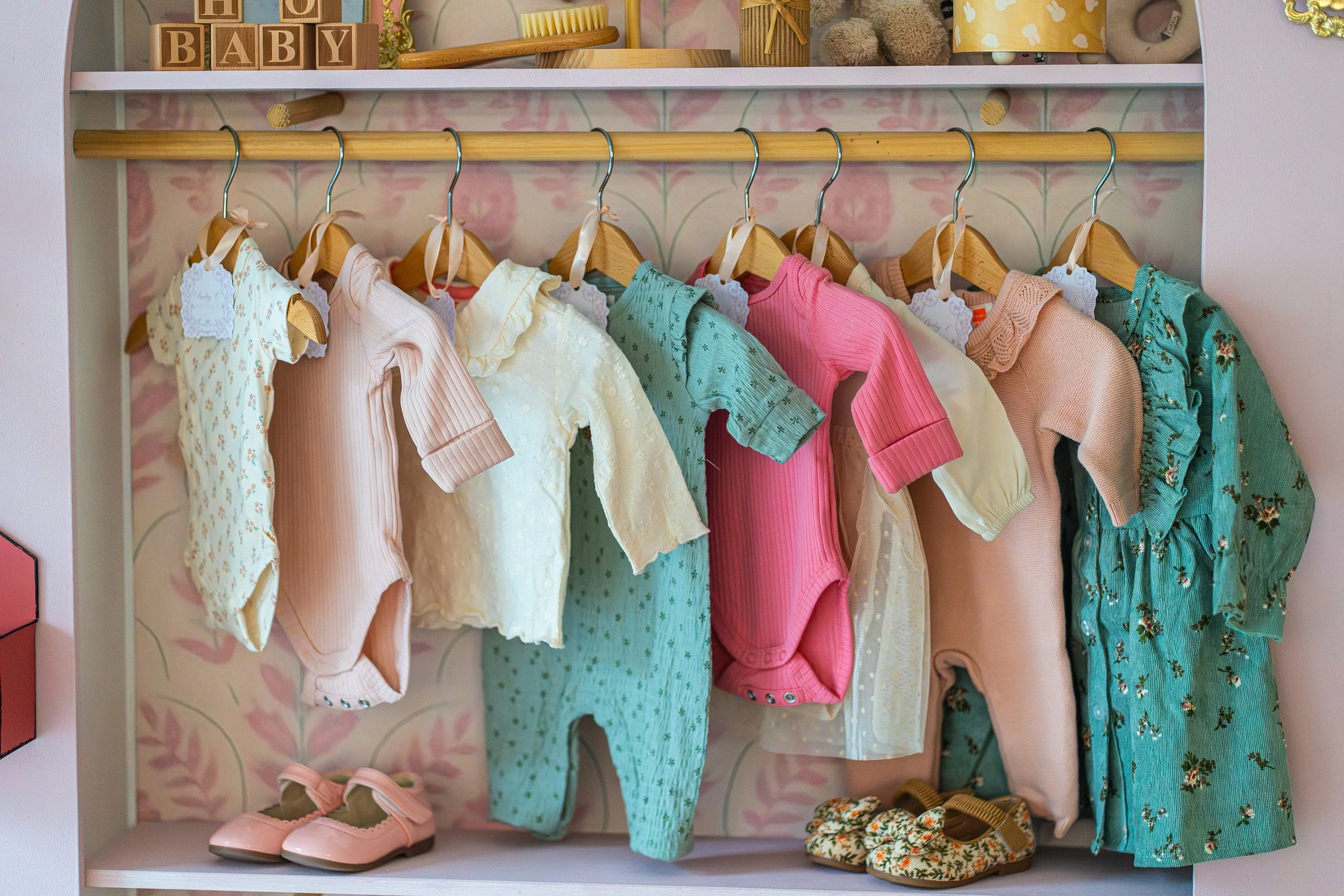 Children's closet with hanging baby clothes in pastel colors, pink shoes at the bottom, and decorative items on a shelf above.