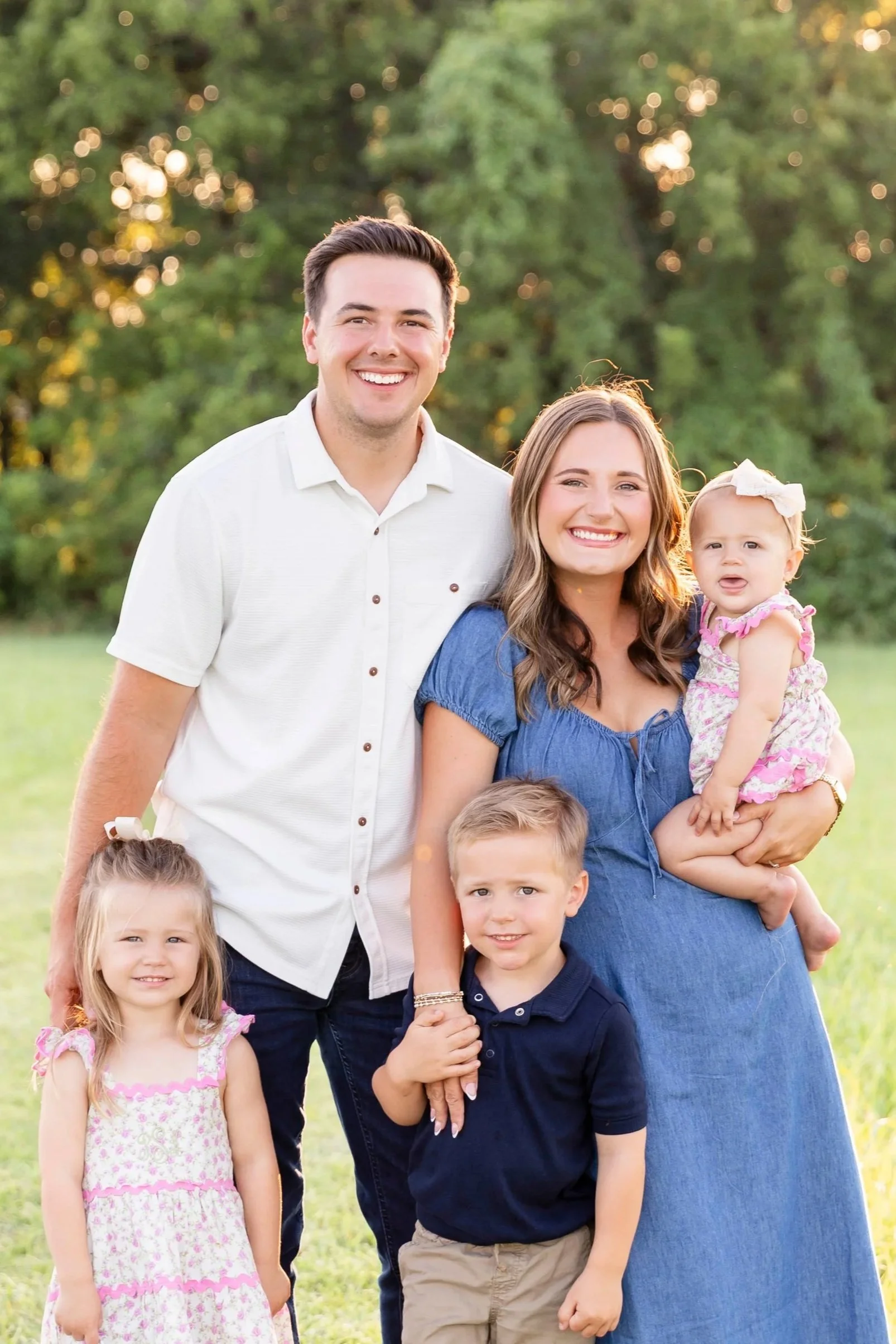 A smiling family of seven standing outdoors in a park during sunset, with trees and greenery in the background. The family includes a man, a woman, and five children, with the woman holding a young girl wearing a pink and white dress.