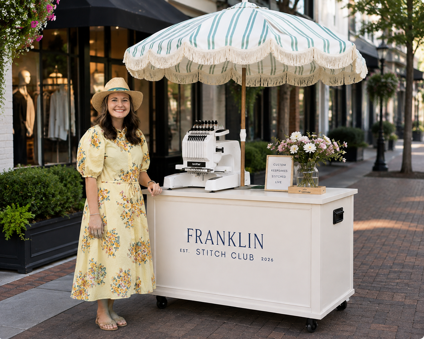 A woman in a yellow floral dress and a wide-brimmed hat stands smiling at a stitch club booth on a city street. The booth has a large umbrella, a vintage sewing machine, a sign that reads 'CUSTOM KEEPSAKES STITCHED LIVE,' and a vase of flowers. The front of the booth says 'FRANKLIN EST. STITCH CLUB 2026.'