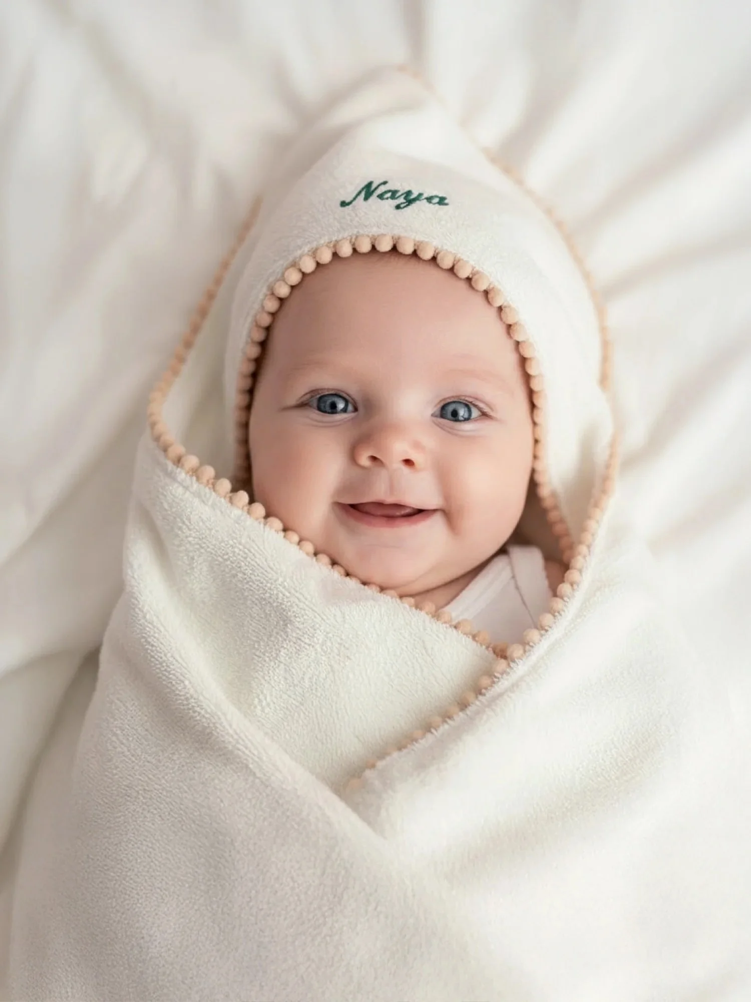 Close-up of a smiling baby wearing a white hoodie with pink beaded trim and the name "Naya" embroidered on the hood.