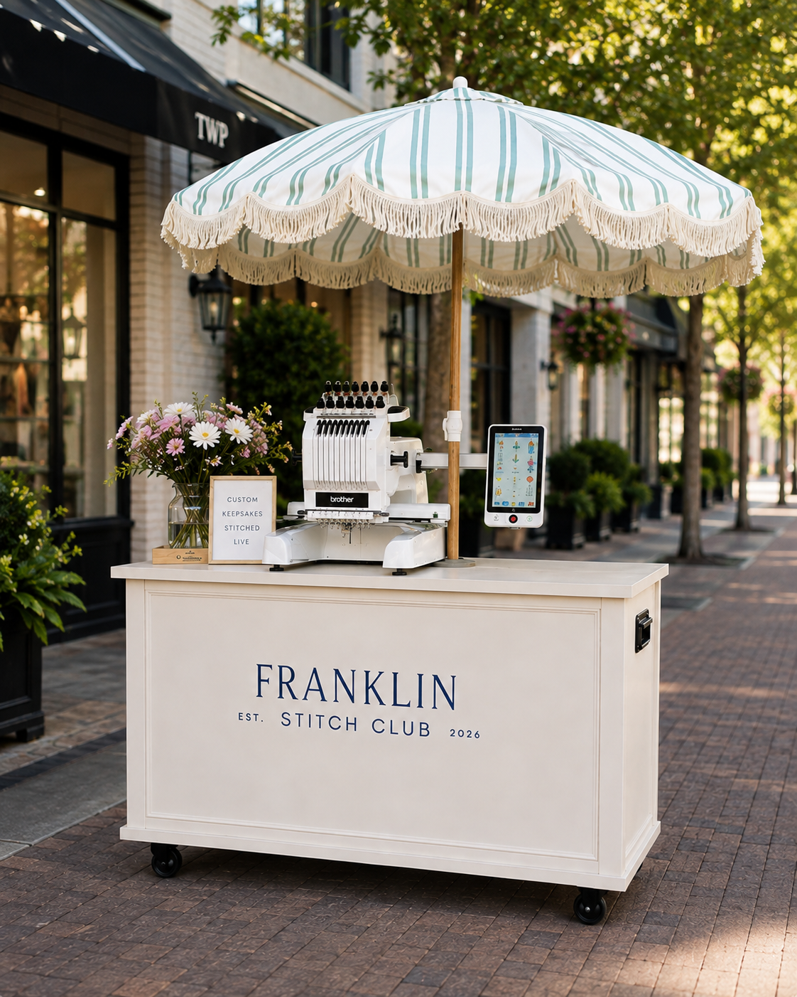 A mobile embroidery shop named Franklin Stitch Club with a white cart, a large striped umbrella, and a embroidery machine, set up on a brick sidewalk, with store fronts, potted plants, and trees in the background.
