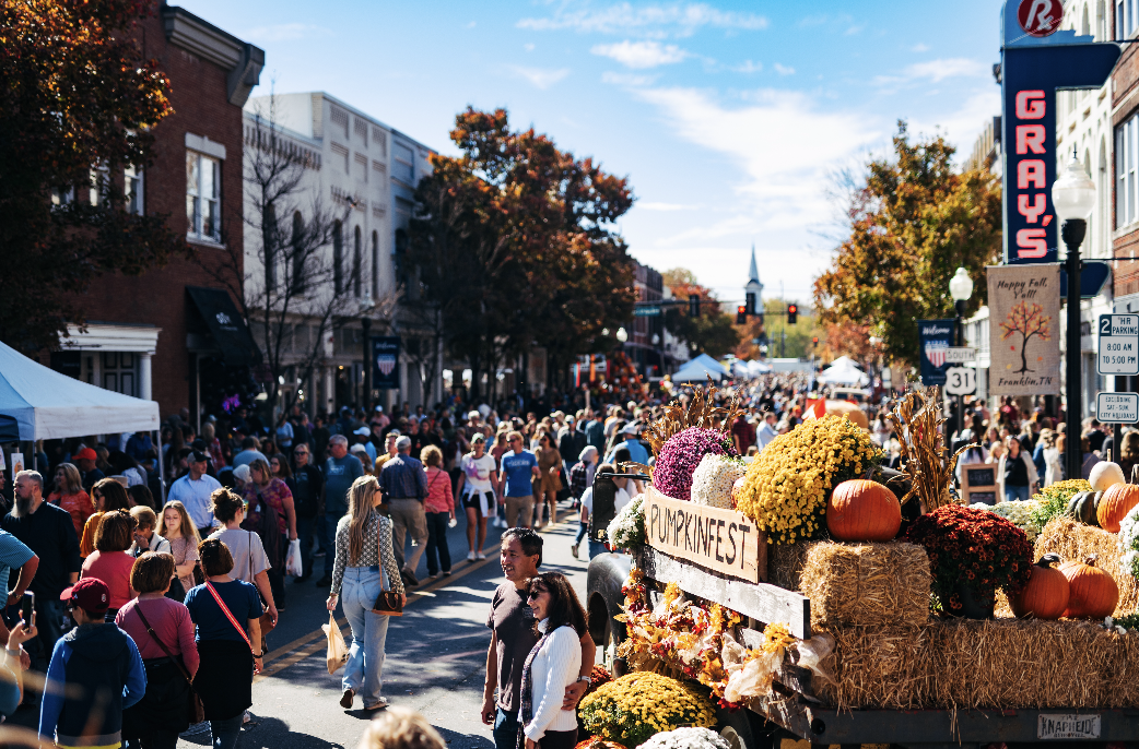 A bustling street fair during fall with a crowd of people walking, colorful autumn trees, and vendor stalls with pumpkins and flowers, including a sign labeled 'PUMPKIN'.