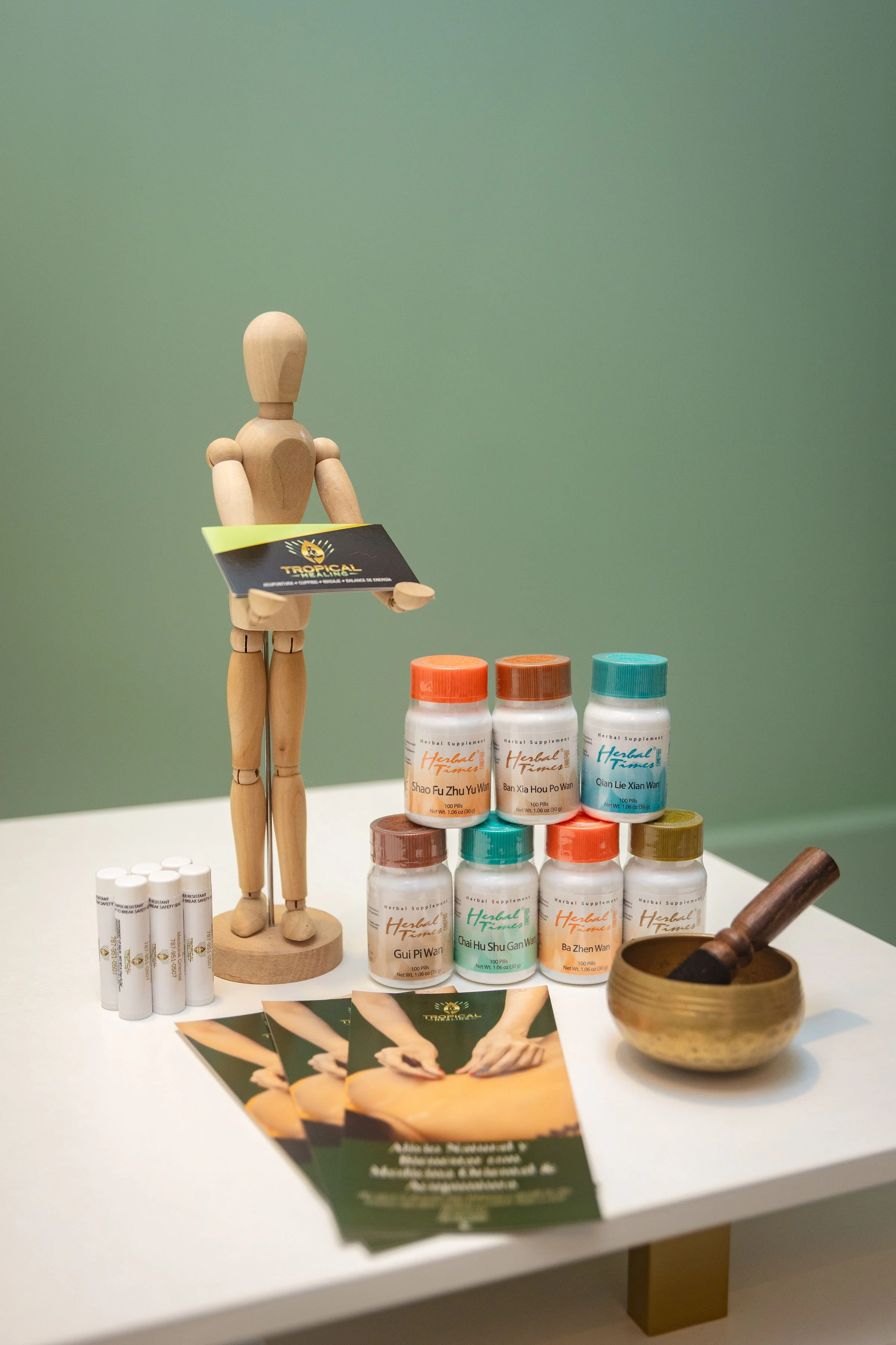 A wooden mannequin holding a flyer, surrounded by bottles of herbal supplements, scattered pamphlets, and a wooden bowl with a scoop, on a white table against a green background.
