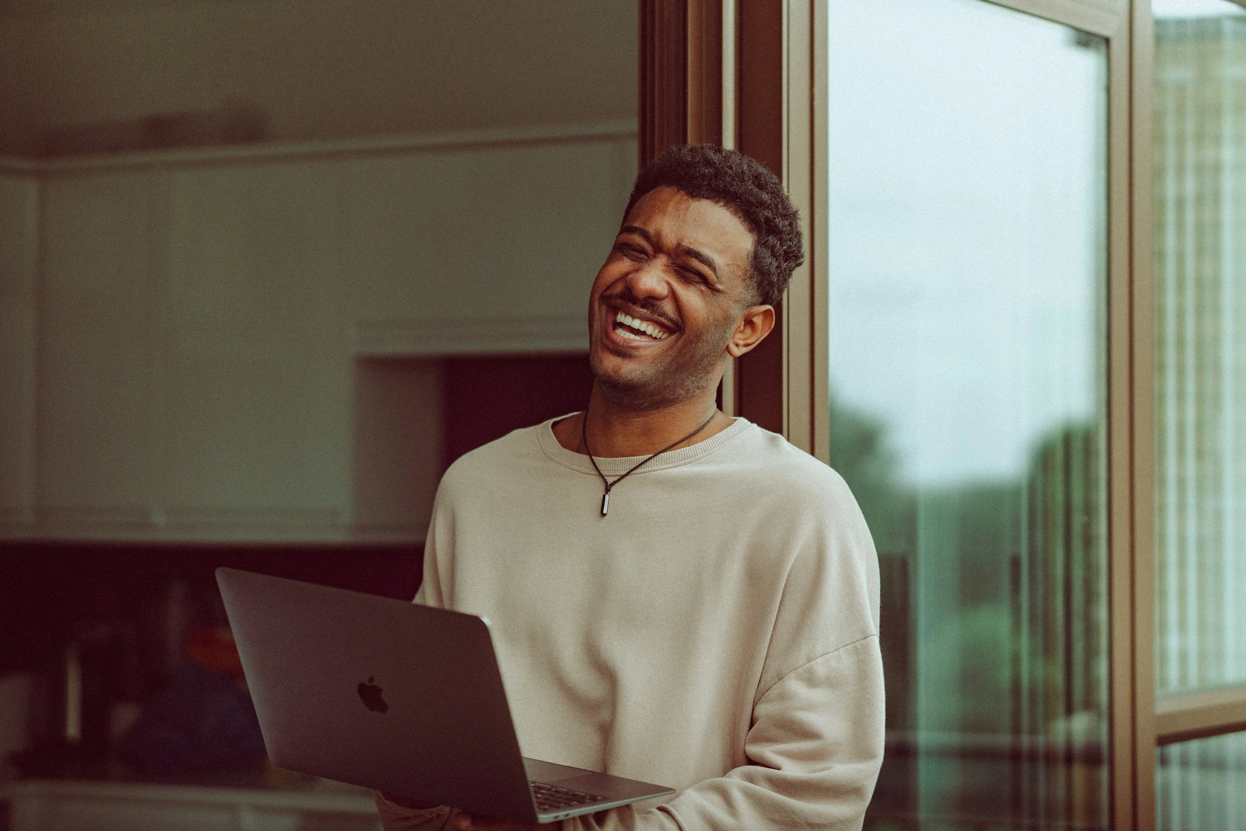 A young man with curly hair and a beard smiling and looking to the side, holding a silver laptop, standing near a wooden-framed glass door or window in a modern interior.