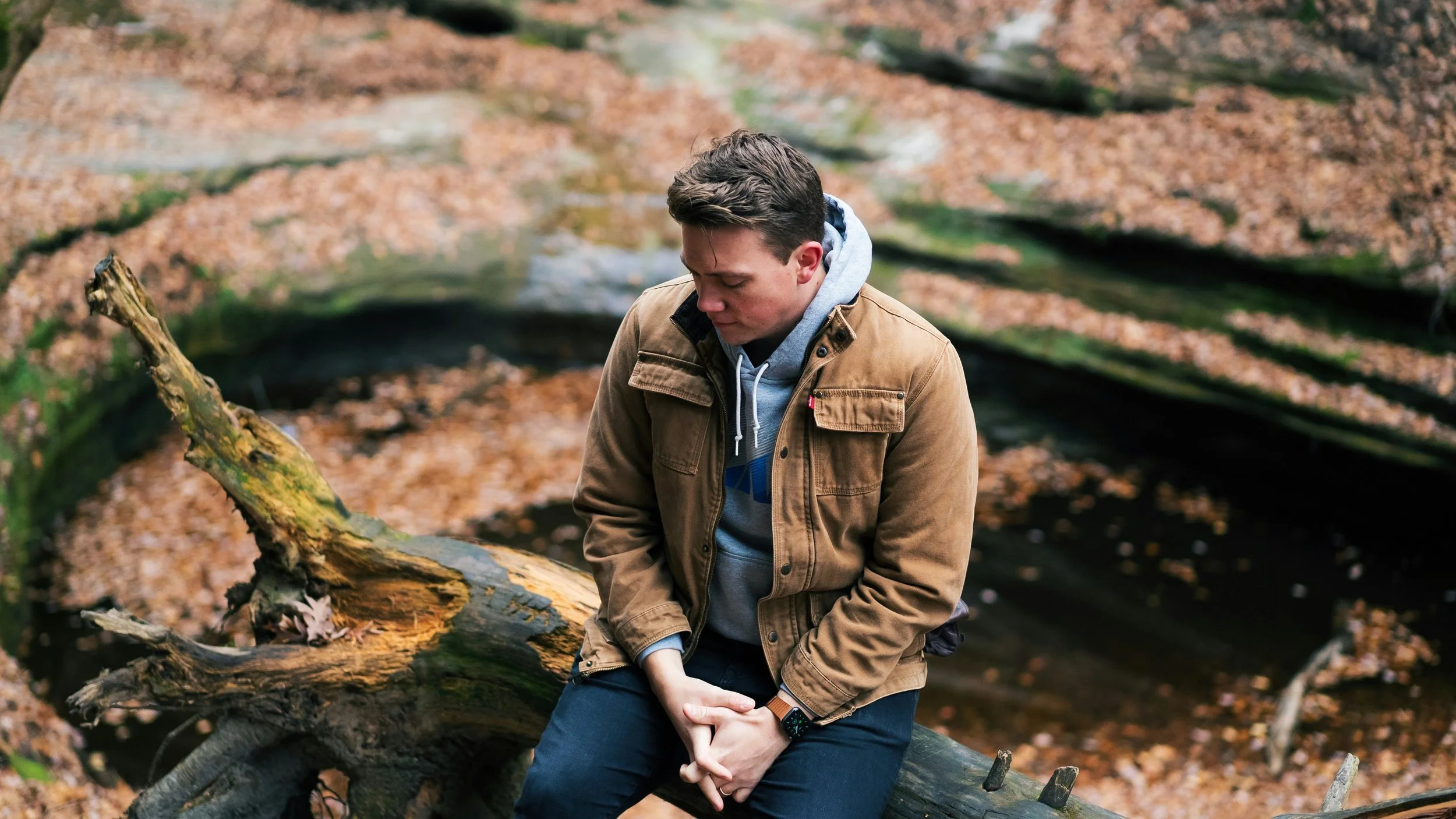 A young man in a tan jacket and gray hoodie sitting on a fallen log in a forest with autumn leaves.