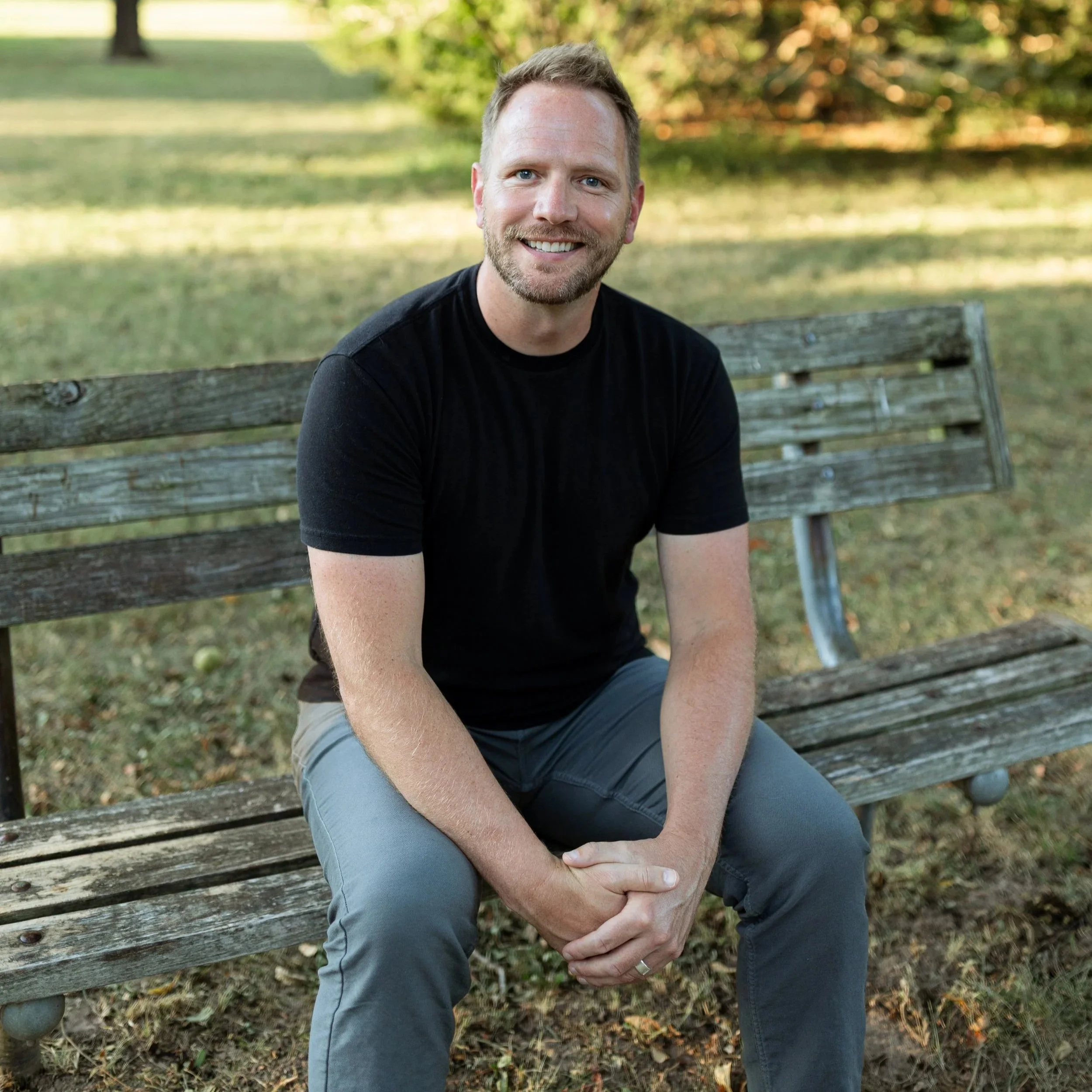 A man sitting on a wooden park bench smiling, outdoors in a green park with trees in the background.