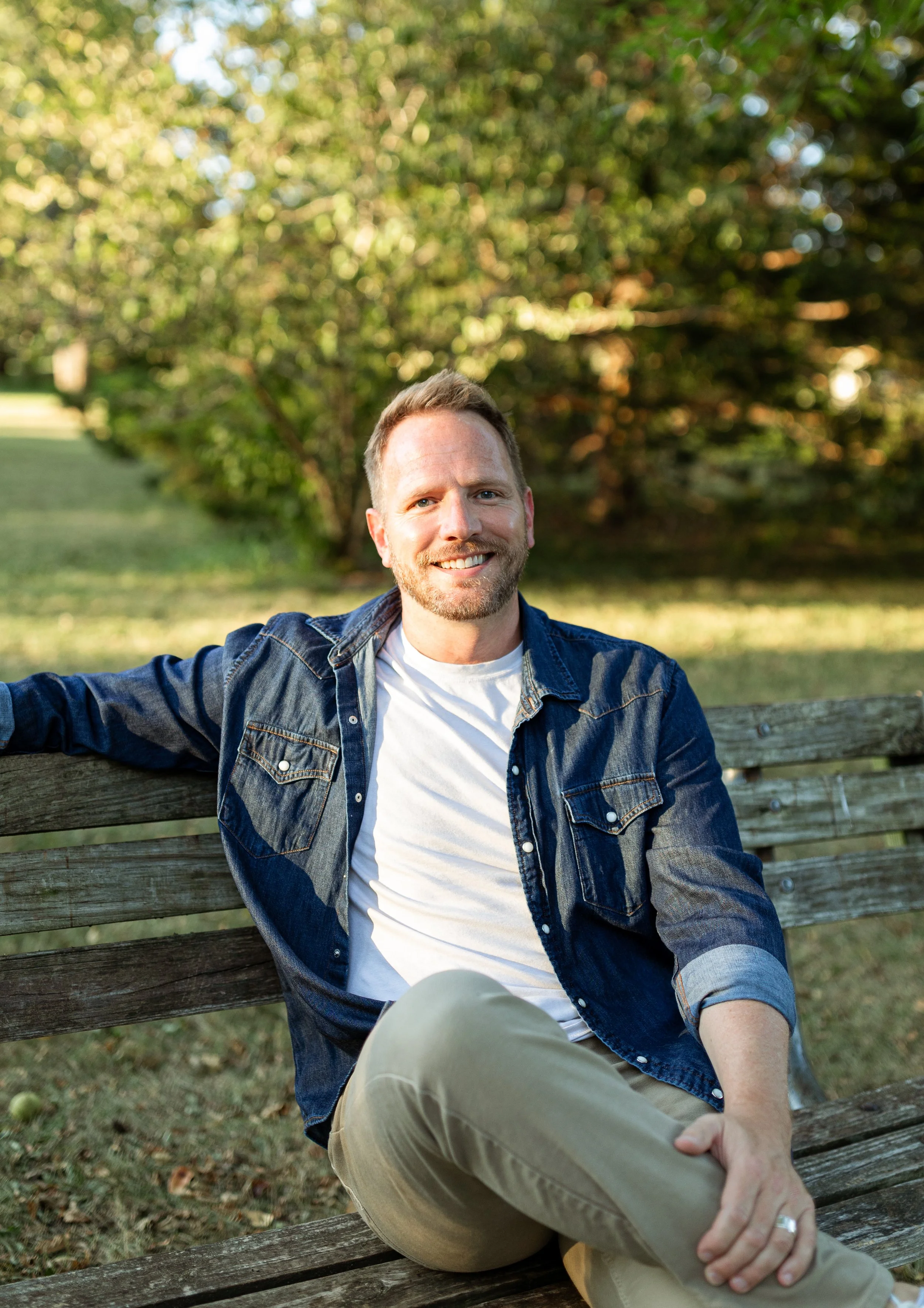 A smiling man sitting on a wooden park bench with greenery and trees in the background, dressed casually in a denim jacket and khaki pants.