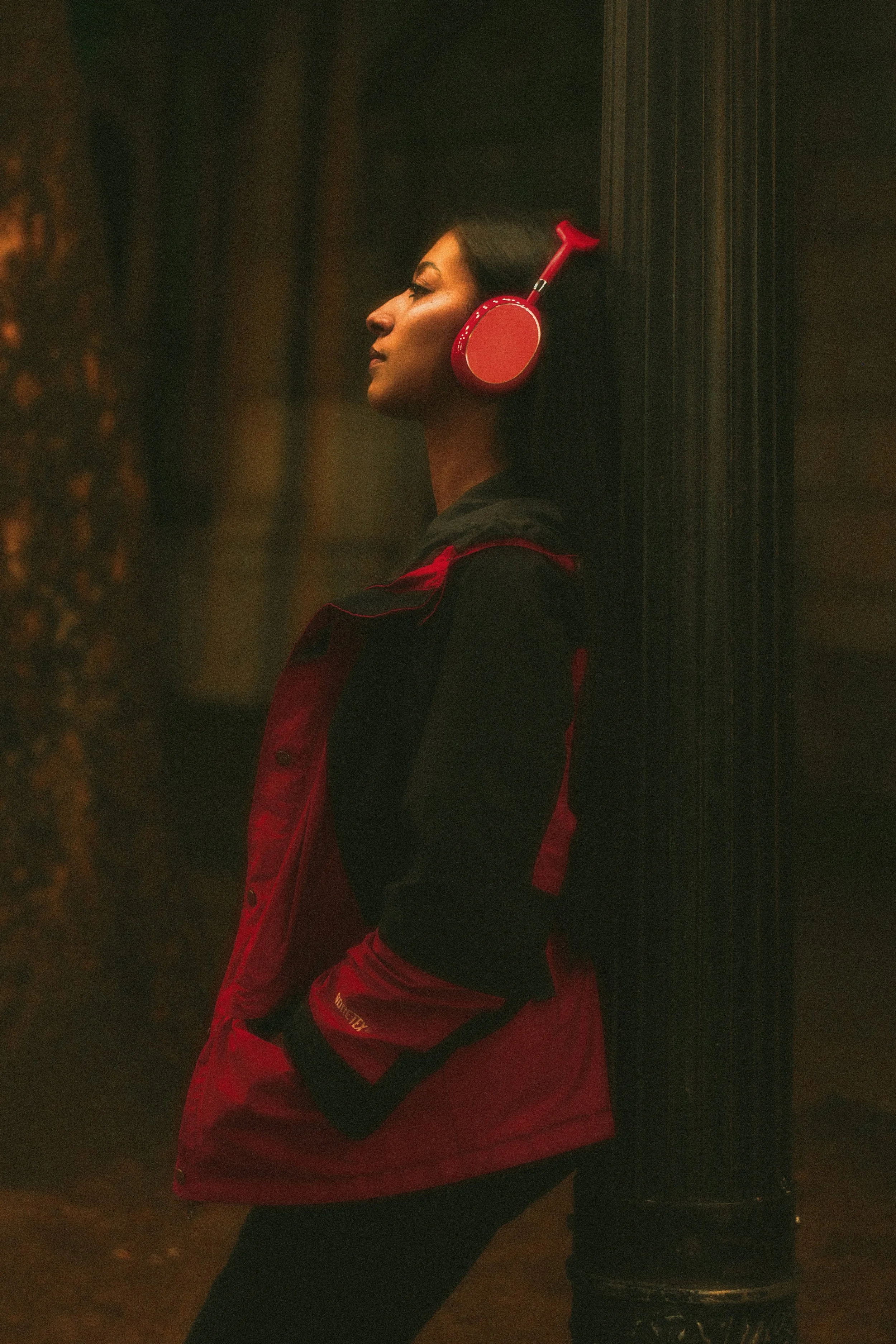 Side view of a woman with dark hair wearing red headphones, a black and red jacket, and red pants, standing against a dark background with warm lighting.