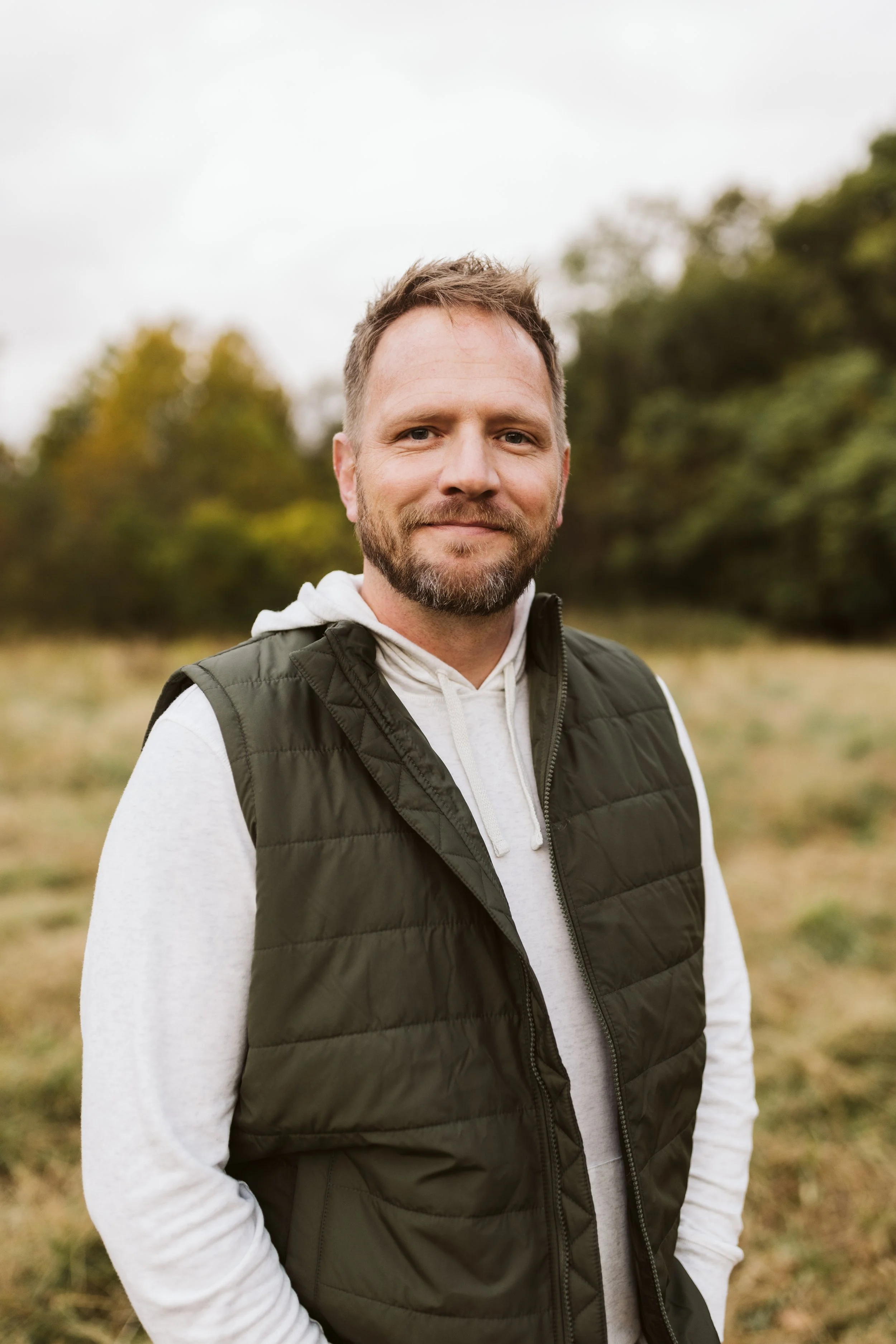 A middle-aged man with short, light brown hair, a beard, and smiling expression, wearing a white hoodie and a dark green puffer vest, standing outdoors in a natural setting with green trees and cloudy sky in the background.
