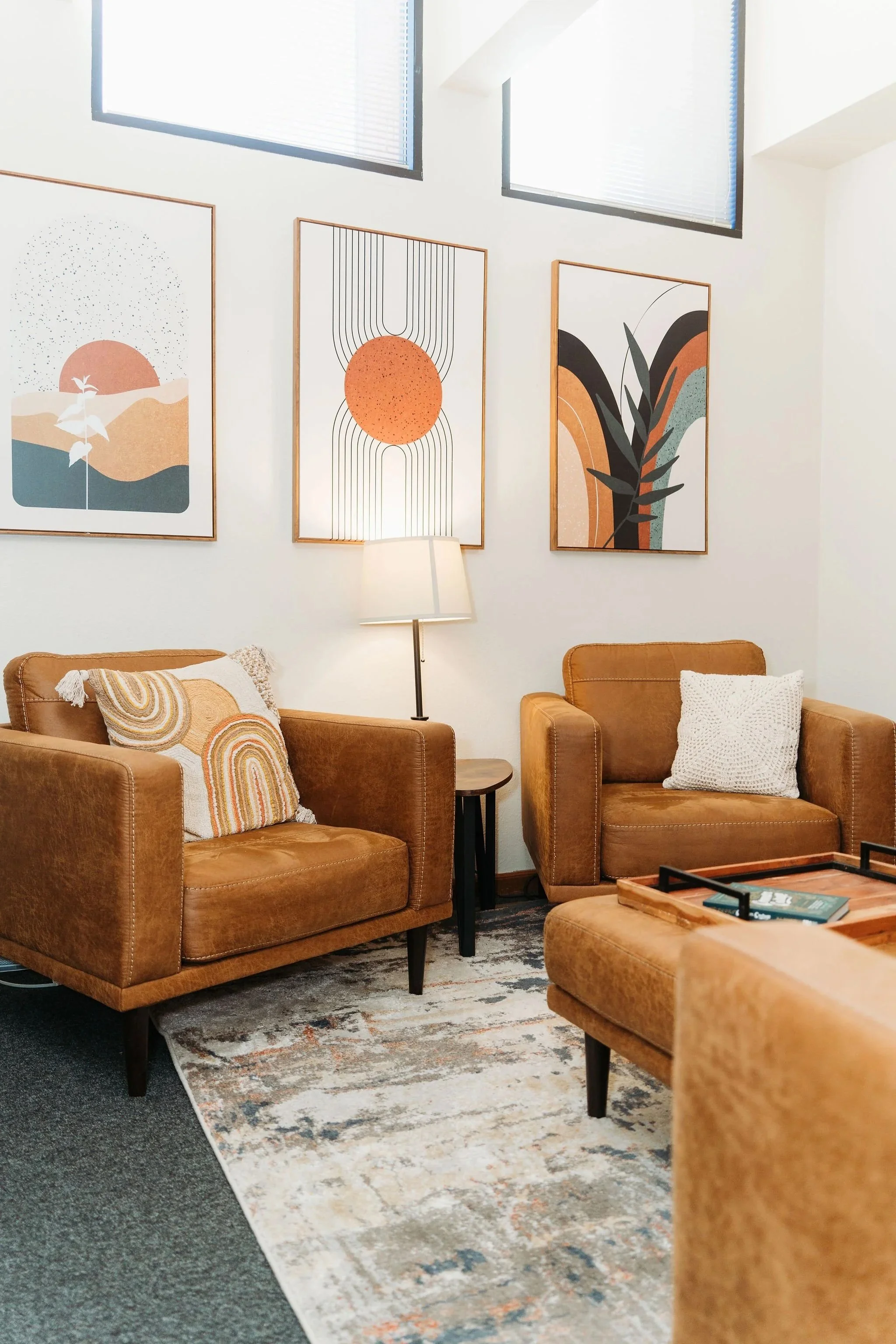 Living room with two tan leather armchairs, a couch, a patterned rug, wall art, a floor lamp, and a wooden coffee table with books.