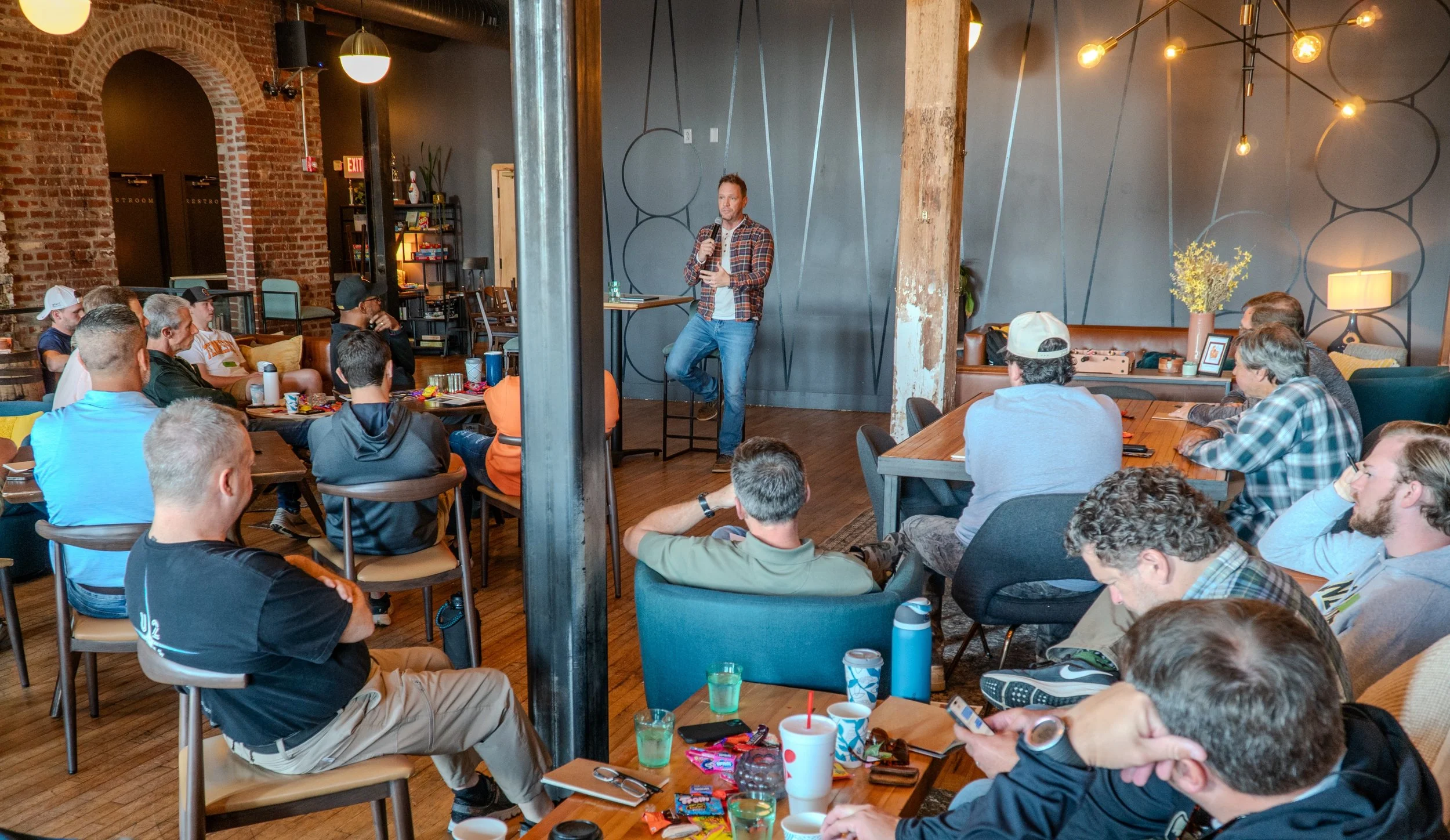 A man in a plaid shirt and jeans is speaking into a microphone on a stage in front of a seated audience in a cozy, modern, brick-walled cafe or event space.
