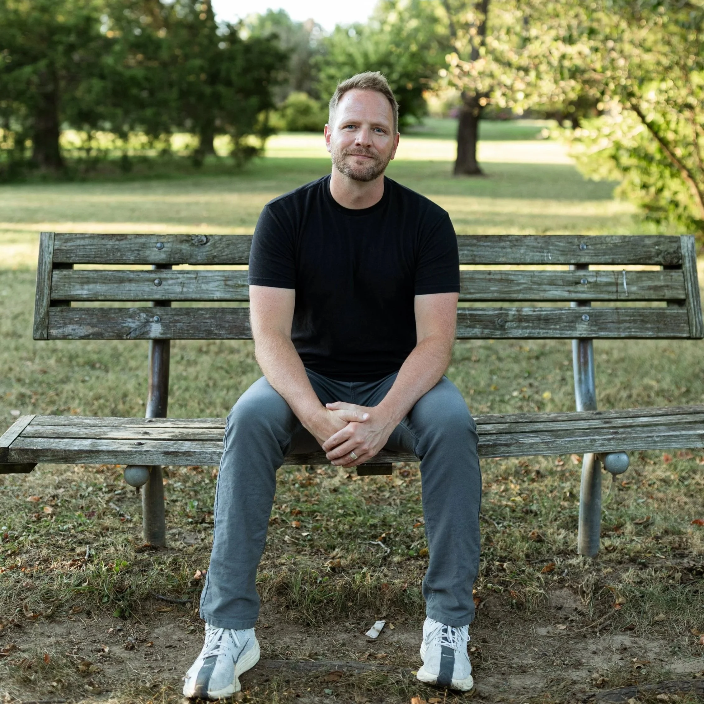 A man sitting on a wooden park bench in a green outdoor area with trees in the background, wearing a black shirt and gray pants, with white sneakers, looking at the camera.