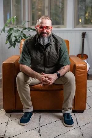 A smiling man with glasses and a graying beard sitting on a brown leather armchair in a brightly lit room with large windows, a plant, and a guitar in the background.