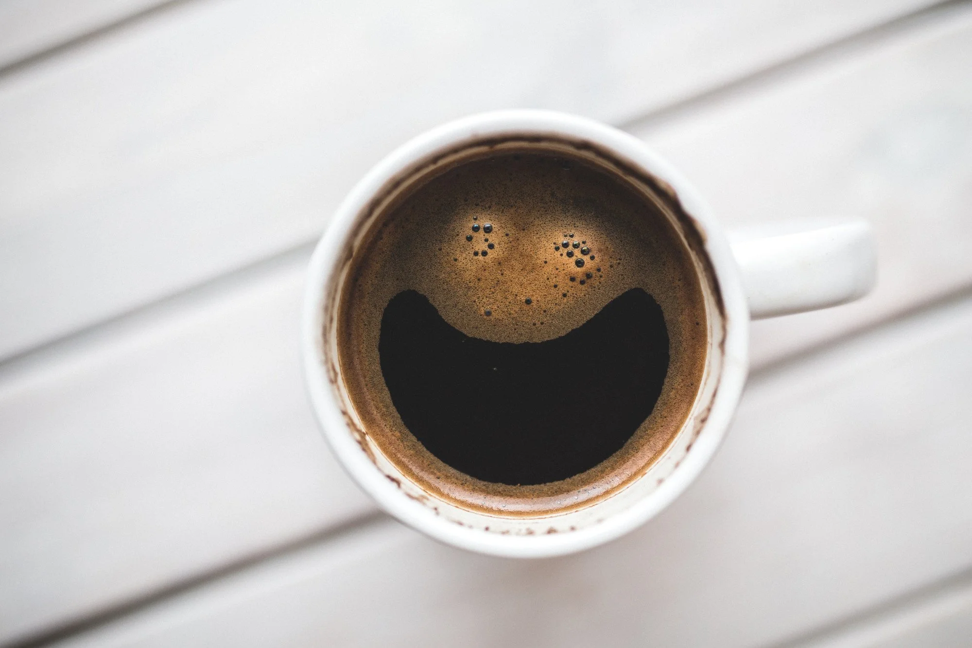 A top-down view of a white mug filled with black coffee on a white wooden surface.