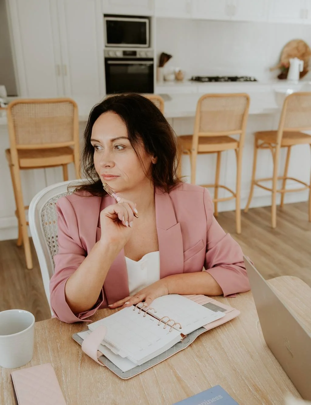 A woman in a pink blazer sitting at a wooden table with a planner, holding a pink pen, in a bright kitchen with white cabinets and bar stools.