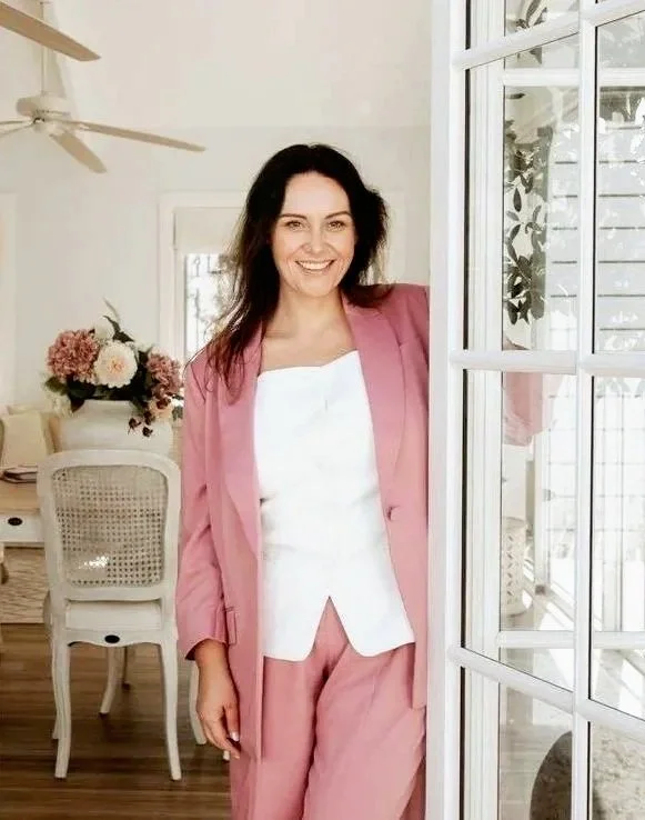 A woman in a pink suit and white top standing near a glass door with a smile in a bright room with floral decor.