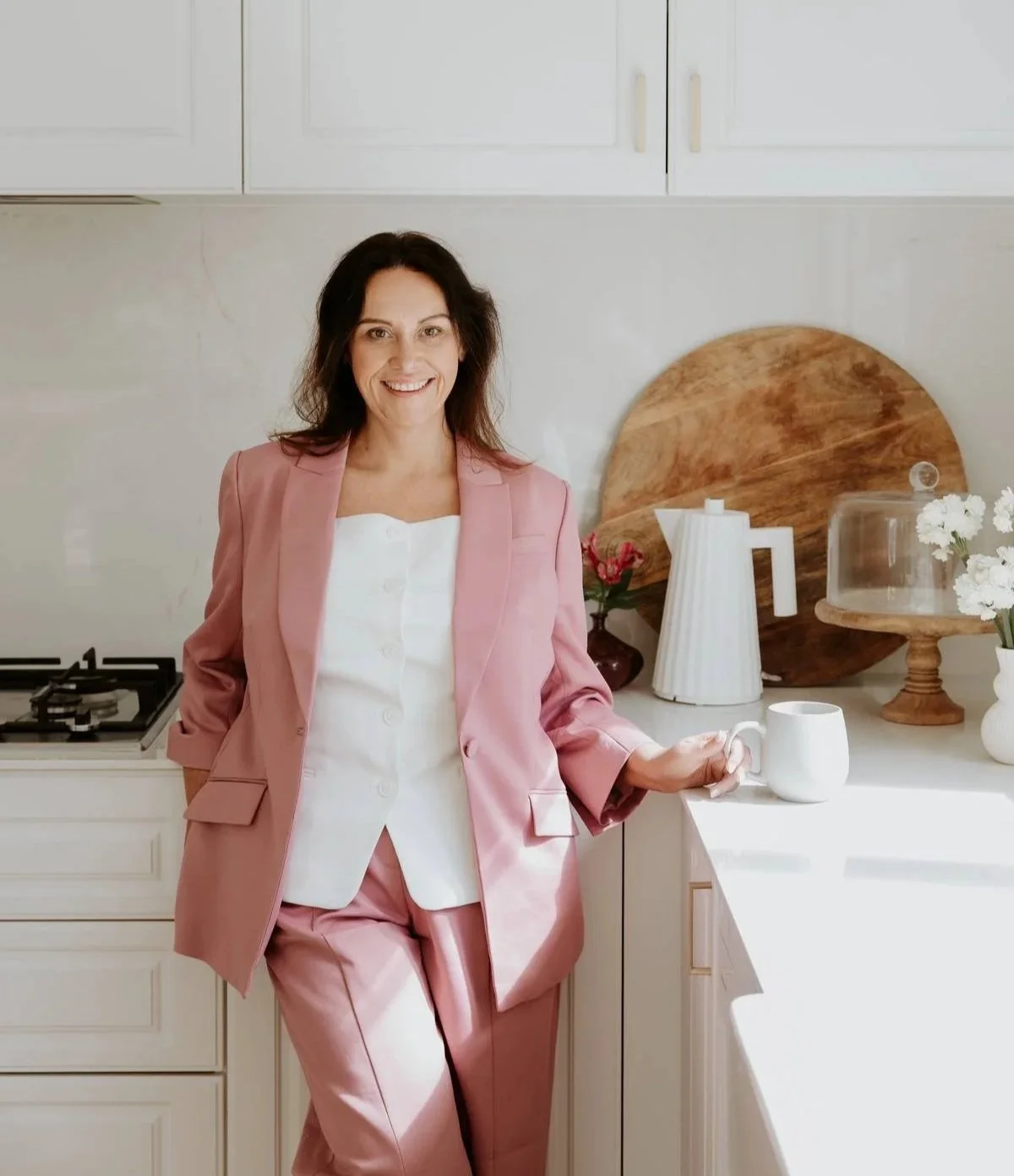 A woman in a pink suit and white top holding a white mug in a bright kitchen with white cabinets, a wooden cutting board, flowers, and a teapot.