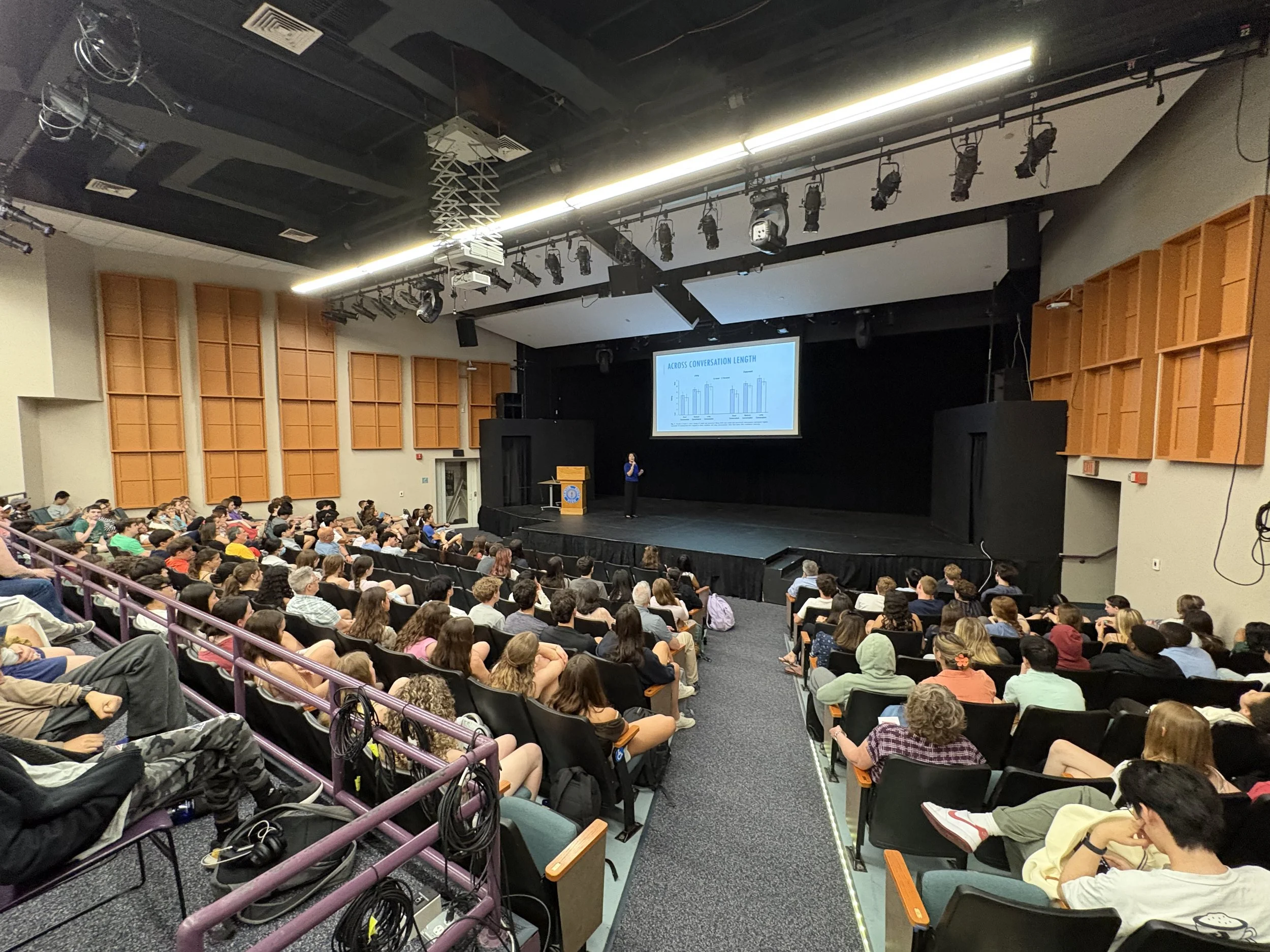 A large auditorium filled with seated audience members watching a presentation. A woman stands on stage near a podium, giving a talk. A large screen behind her displays a graph titled 'Across Conversation Length'. The auditorium has black ceiling with multiple stage lights and audio equipment hanging from it, along with orange acoustic panels on the walls.