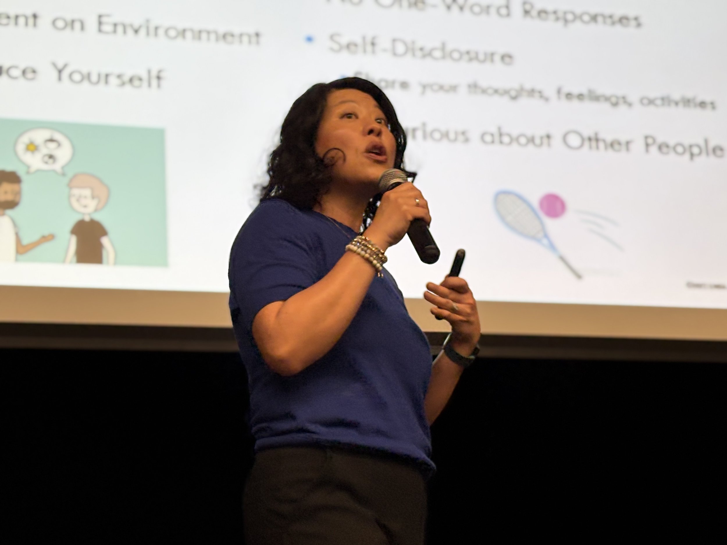 A woman with dark, curly hair holding a microphone, giving a presentation in front of a slide with text about environmental considerations.