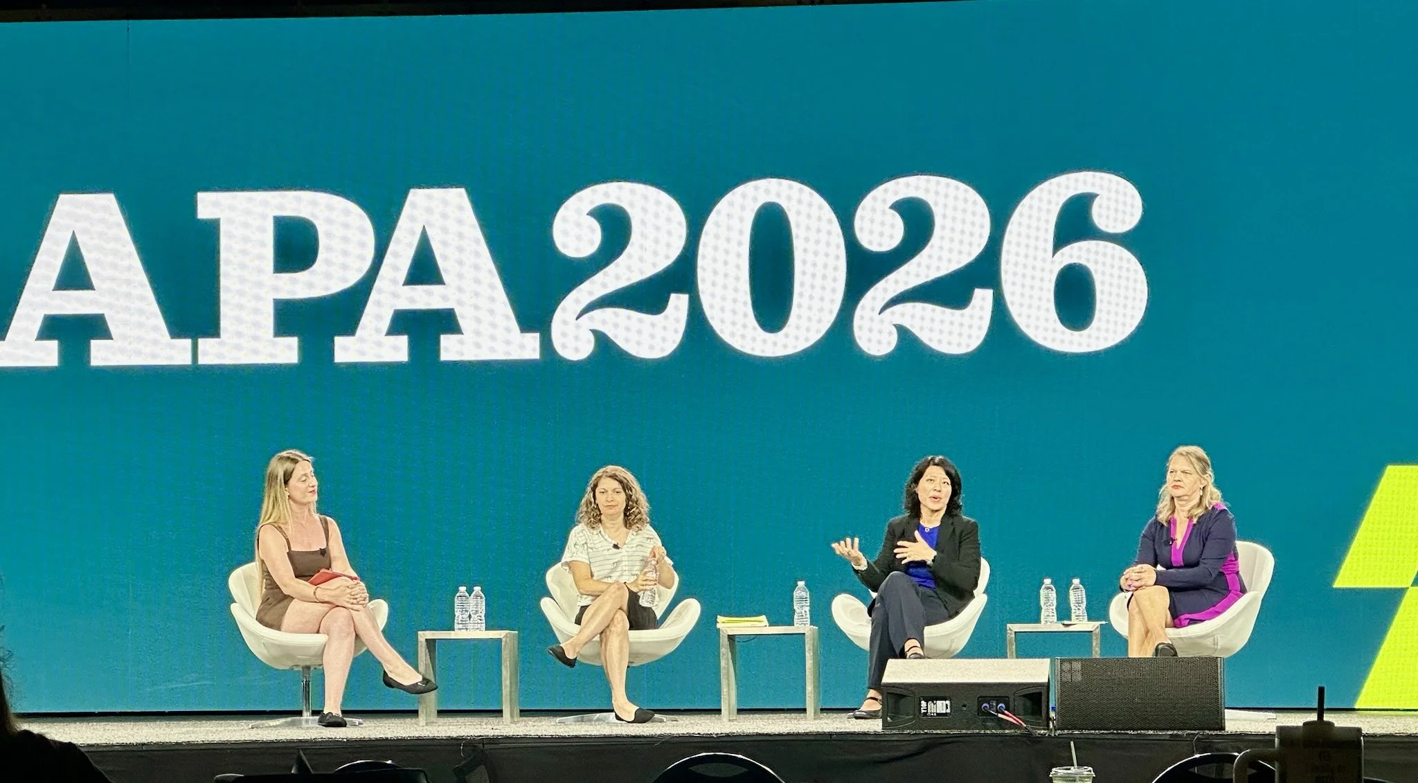 Four women seated on stage at a panel discussion with a large blue background displaying the text 'APA 2026' and yellow geometric designs.