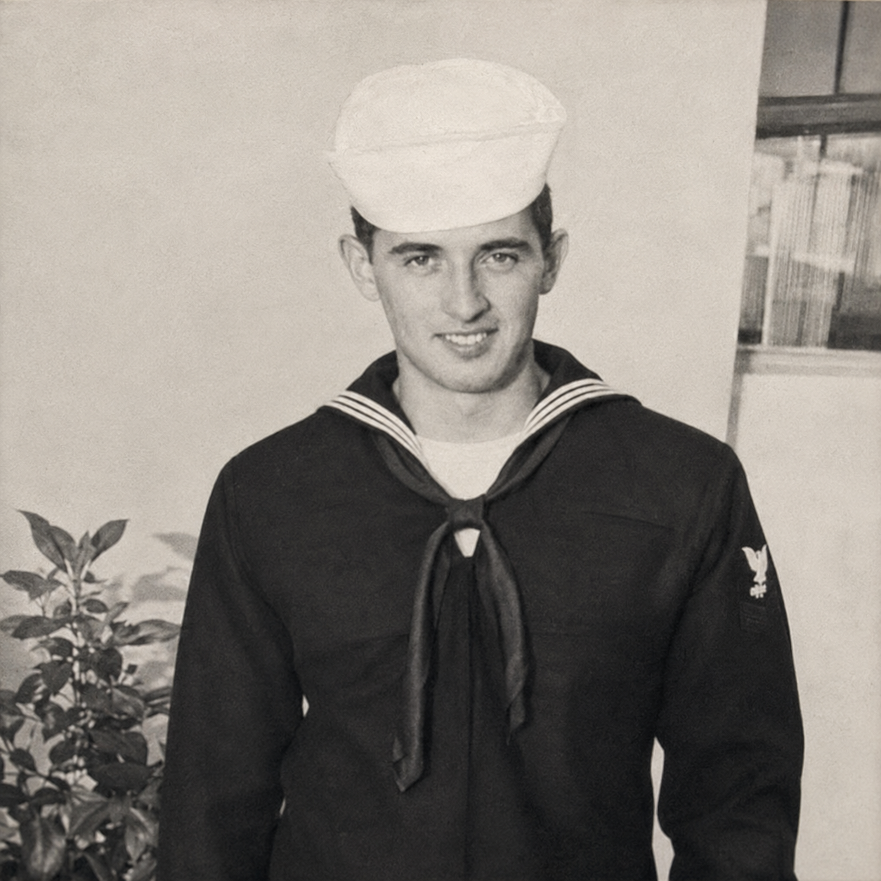Black and white photo of a young man in a navy sailor uniform wearing a white sailor hat, standing indoors near a wall with a window and a plant.