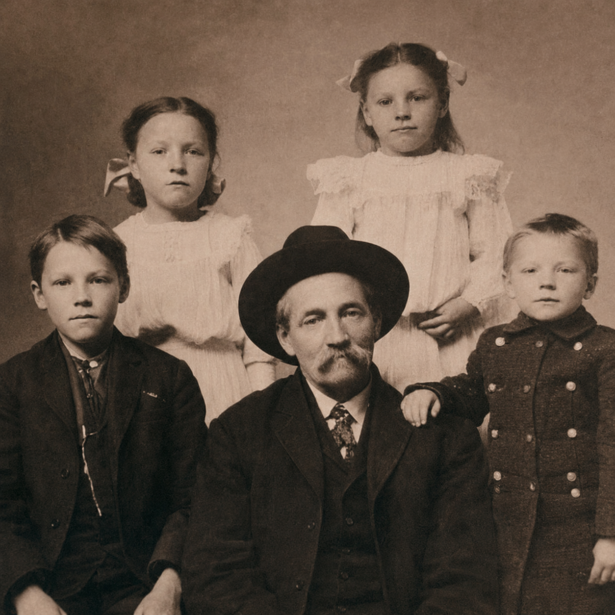 A sepia-toned photograph of an older man with a beard and mustache, wearing a wide-brimmed hat, surrounded by four young children. The children are dressed in vintage clothing, some with bows and buttons, with two girls standing behind and two boys seated beside him, one with his hand on the man's shoulder.