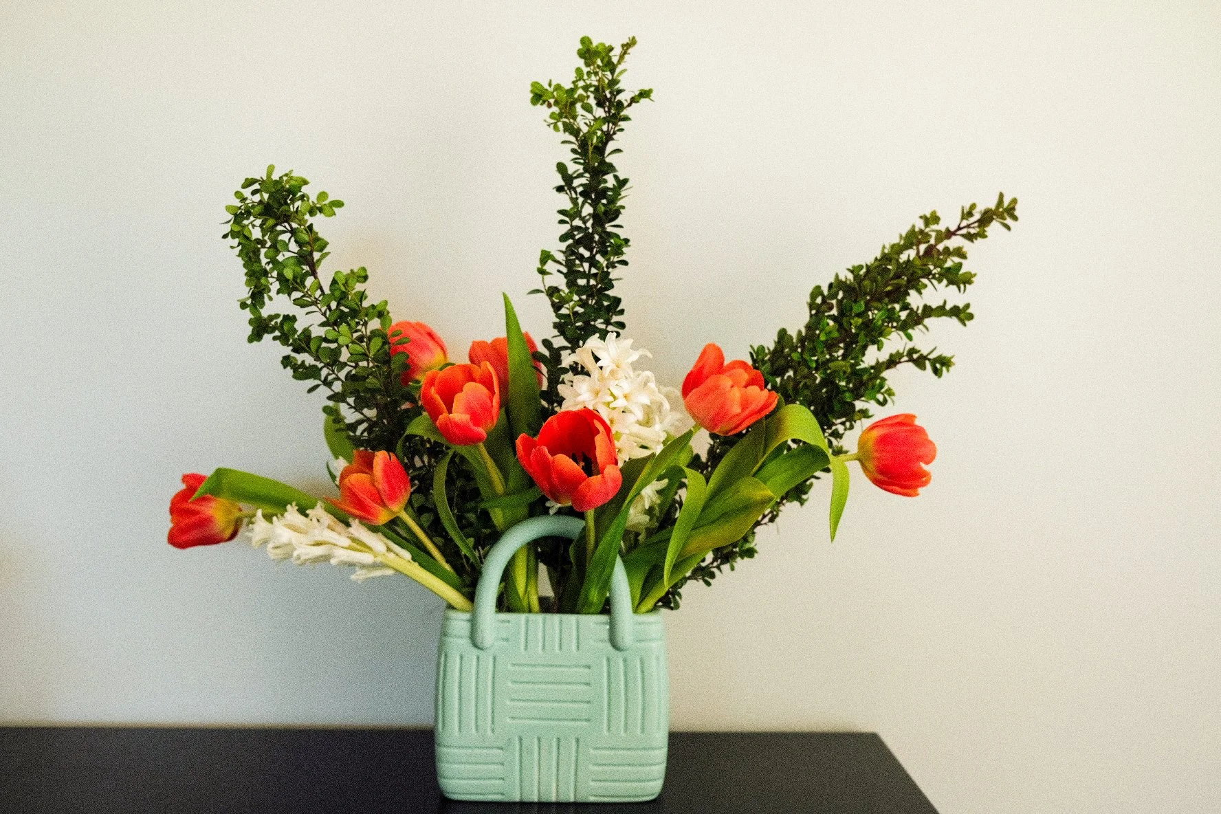 A bouquet of orange tulips and white hyacinths with green foliage in a textured light green vase on a dark surface against a light background.
