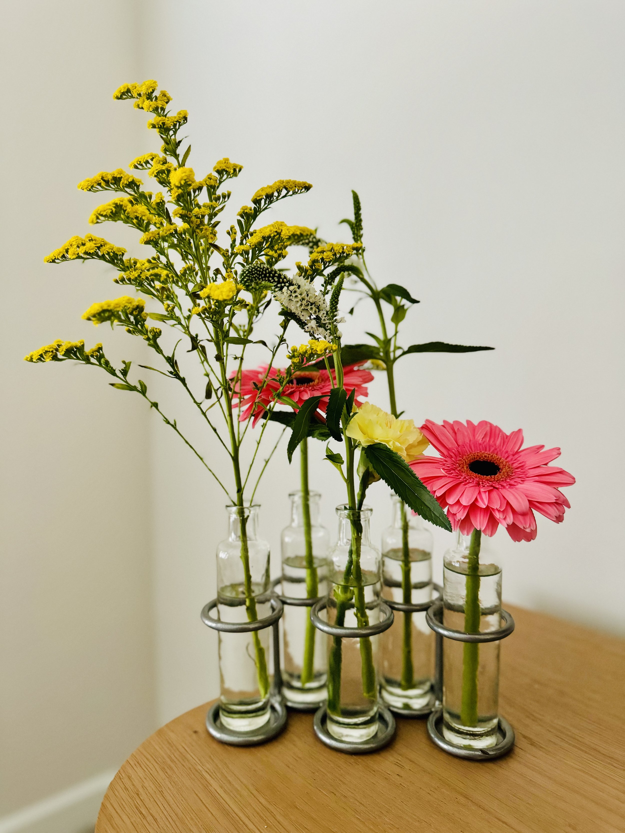 Several small glass test tubes with metal holders containing various flowers placed on a wooden surface against a plain background.