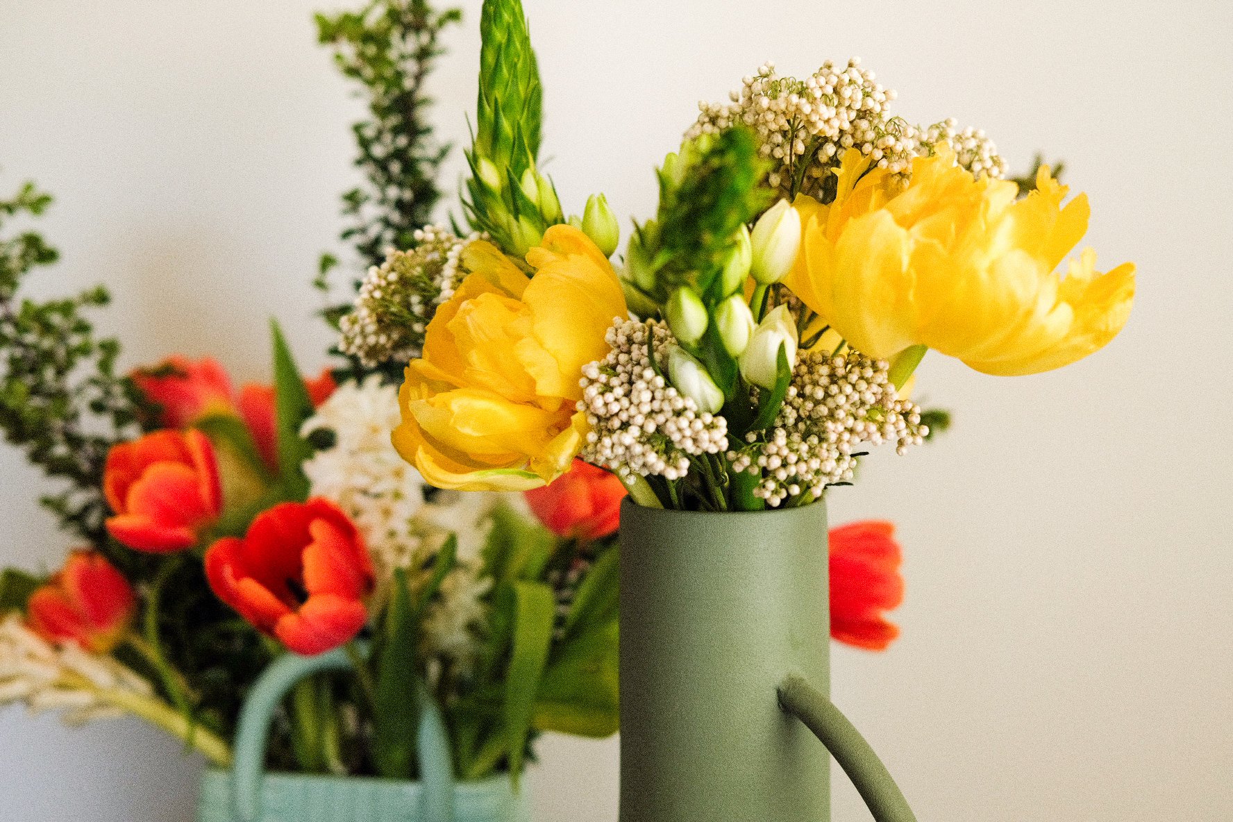 Close-up of a bouquet of flowers in a green vase featuring yellow flowers, white buds, and baby's breath, with additional floral arrangements blurred in the background.