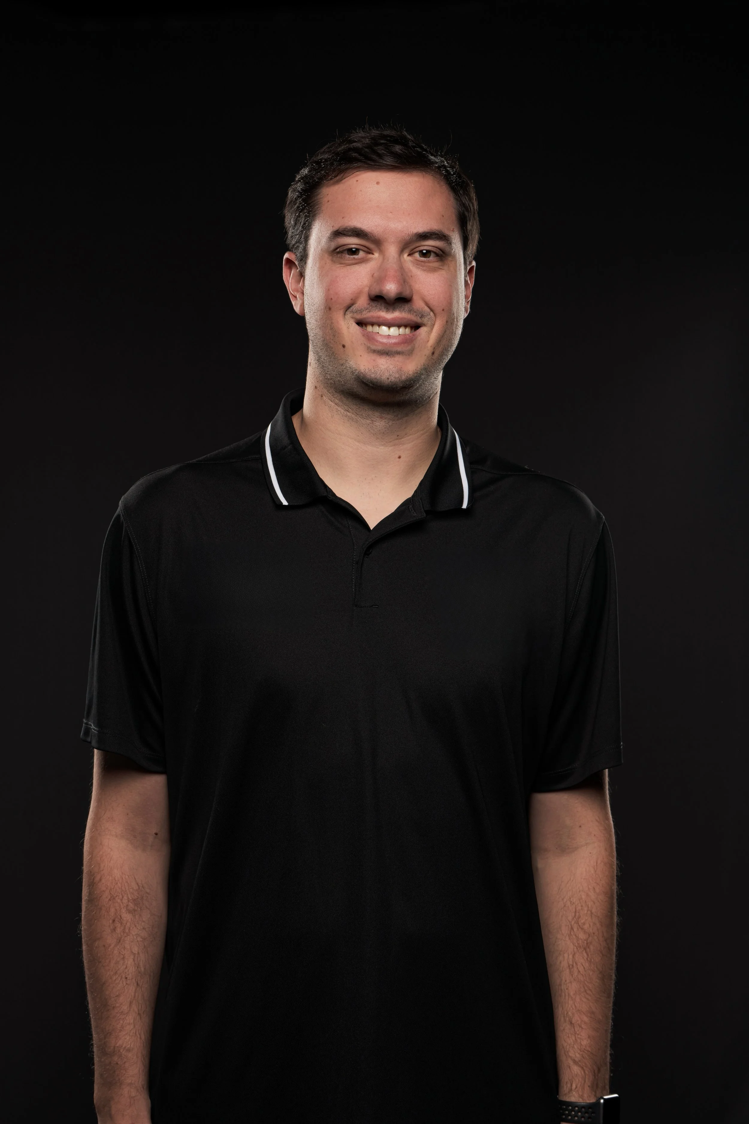 A picture of Sam Davis; a man with short dark hair, light skin, and a slight smile, wearing a black polo shirt, against a dark background.