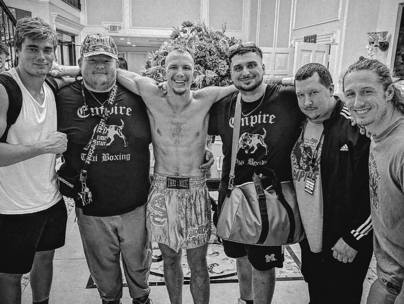 A group of seven men, one shirtless and wearing boxing shorts, poses together indoors in front of a floral arrangement. They are smiling and have their arms around each other.