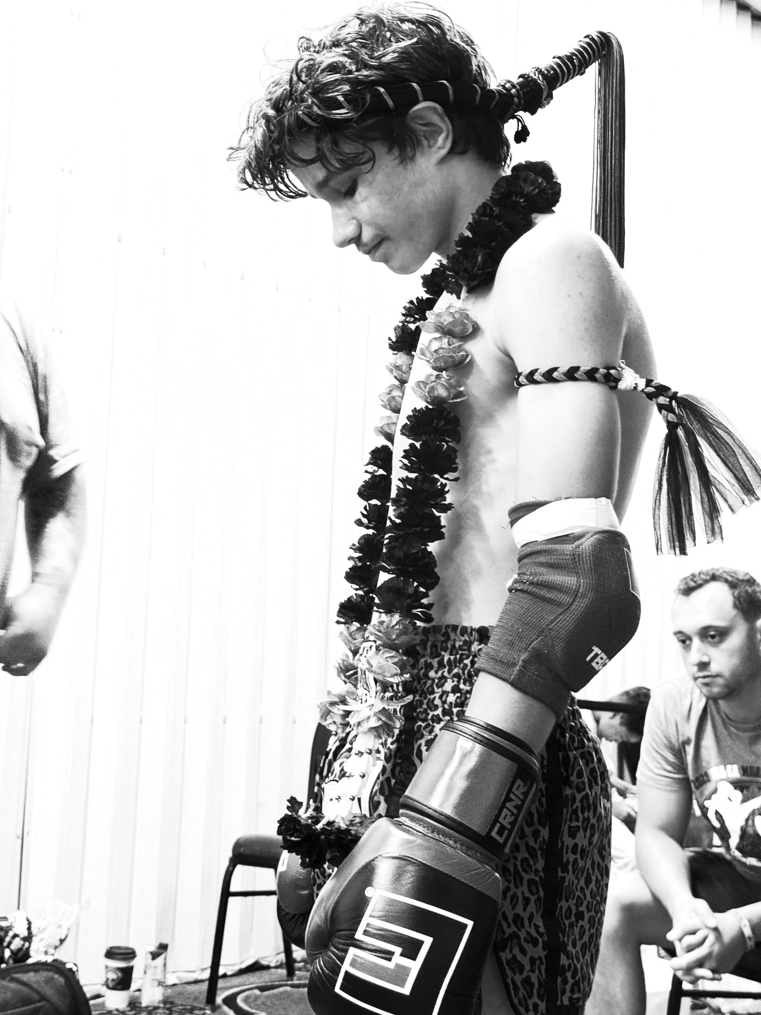 A young person with curly hair wearing a headband and a lei, practicing boxing with gloves on, in a room with a wooden wall. There is a man sitting nearby, holding hands with someone out of frame. The room has various items including a chair, a rug, and coffee cups.