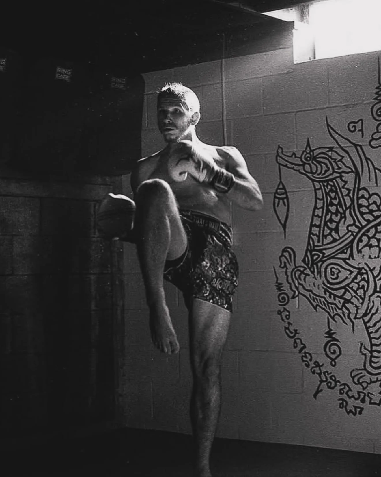 A shirtless man with a mohawk hairstyle practicing Muay Thai or kickboxing poses in a gym with traditional artwork of a Thai mythological creature on the wall.