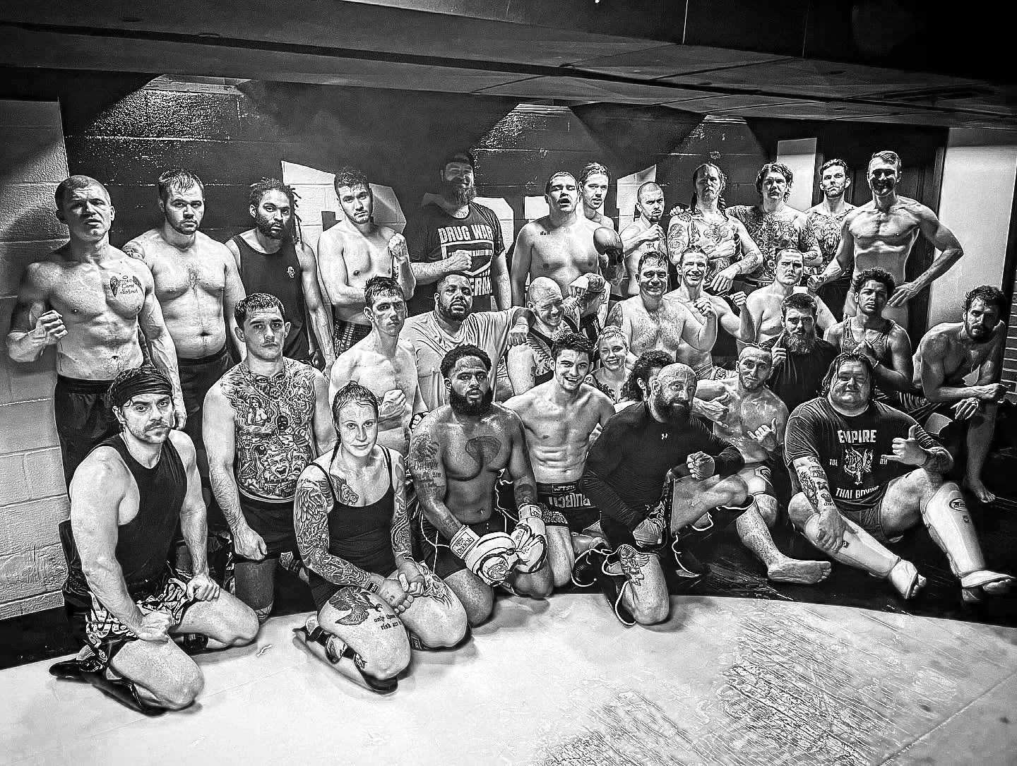 Group of mixed martial arts fighters in a gym, many with tattoos, some wearing gloves and shorts, posing for a group photo.
